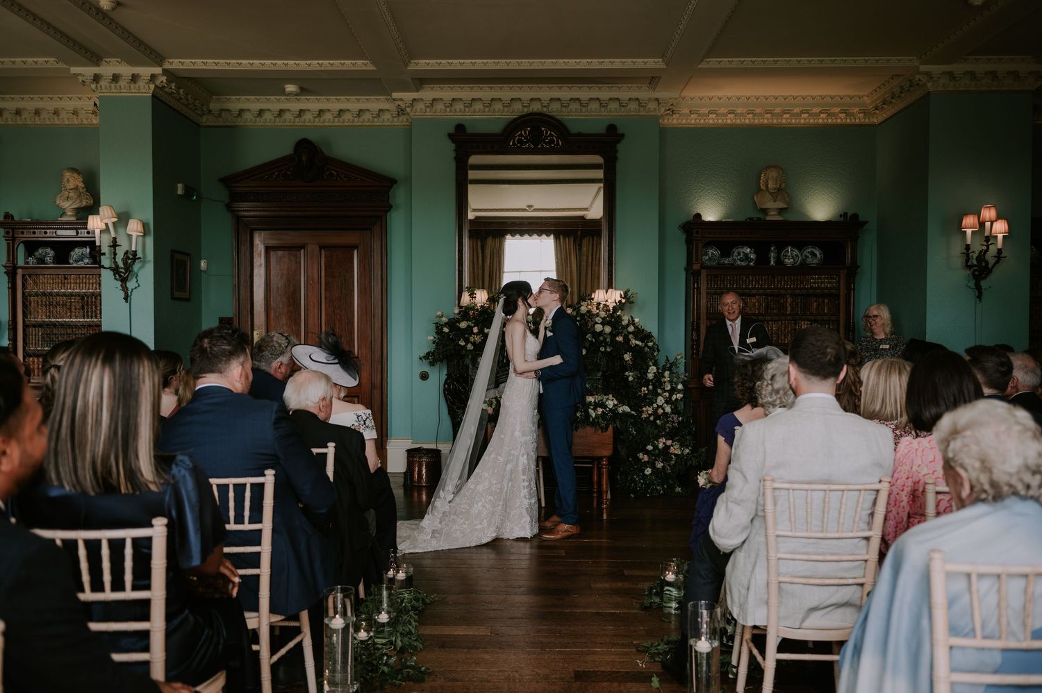 Wedding ceremony guests watch as couple walks down the aisle in an elegant venue with teal walls and ornate details.