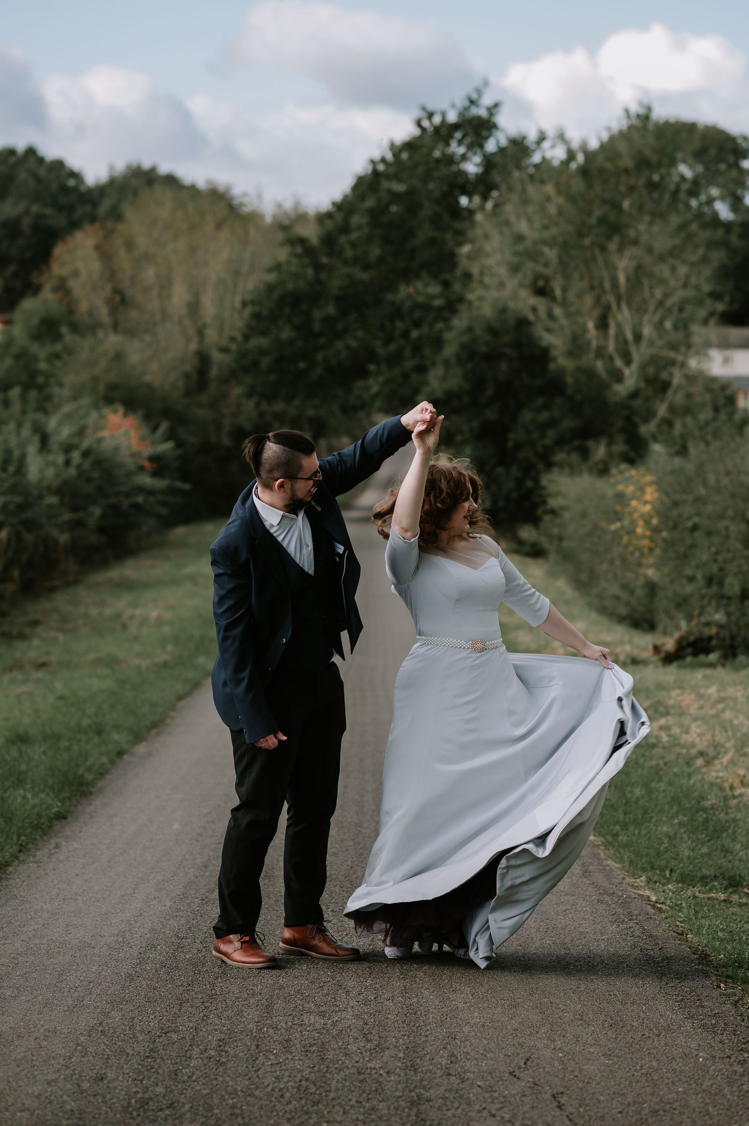 A bride in a flowing white dress twirls on a path while dancing with her partner in a navy suit amid scenic greenery.