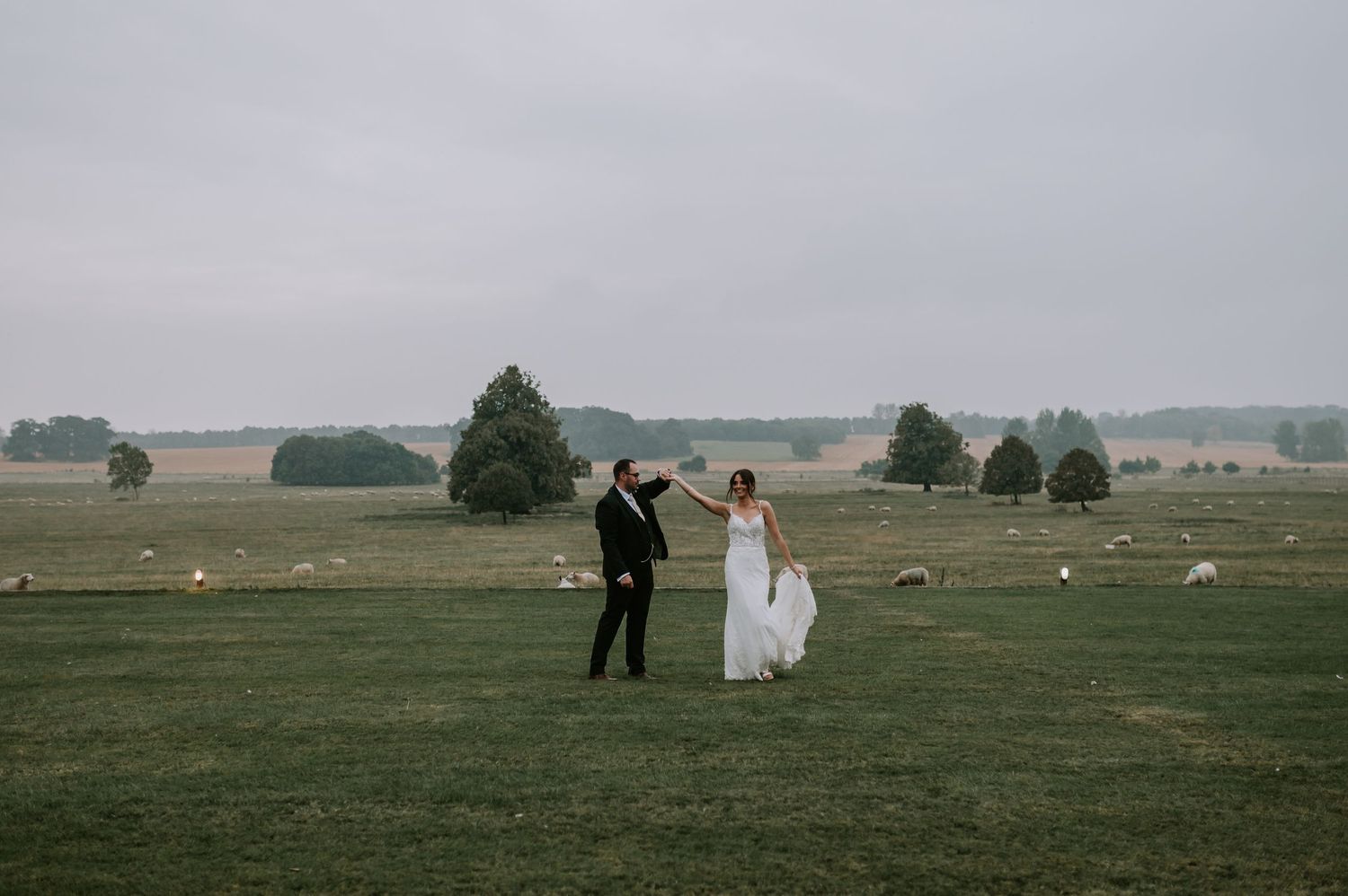 A couple in wedding attire walks hand in hand across a misty green meadow with scattered trees in the distance.