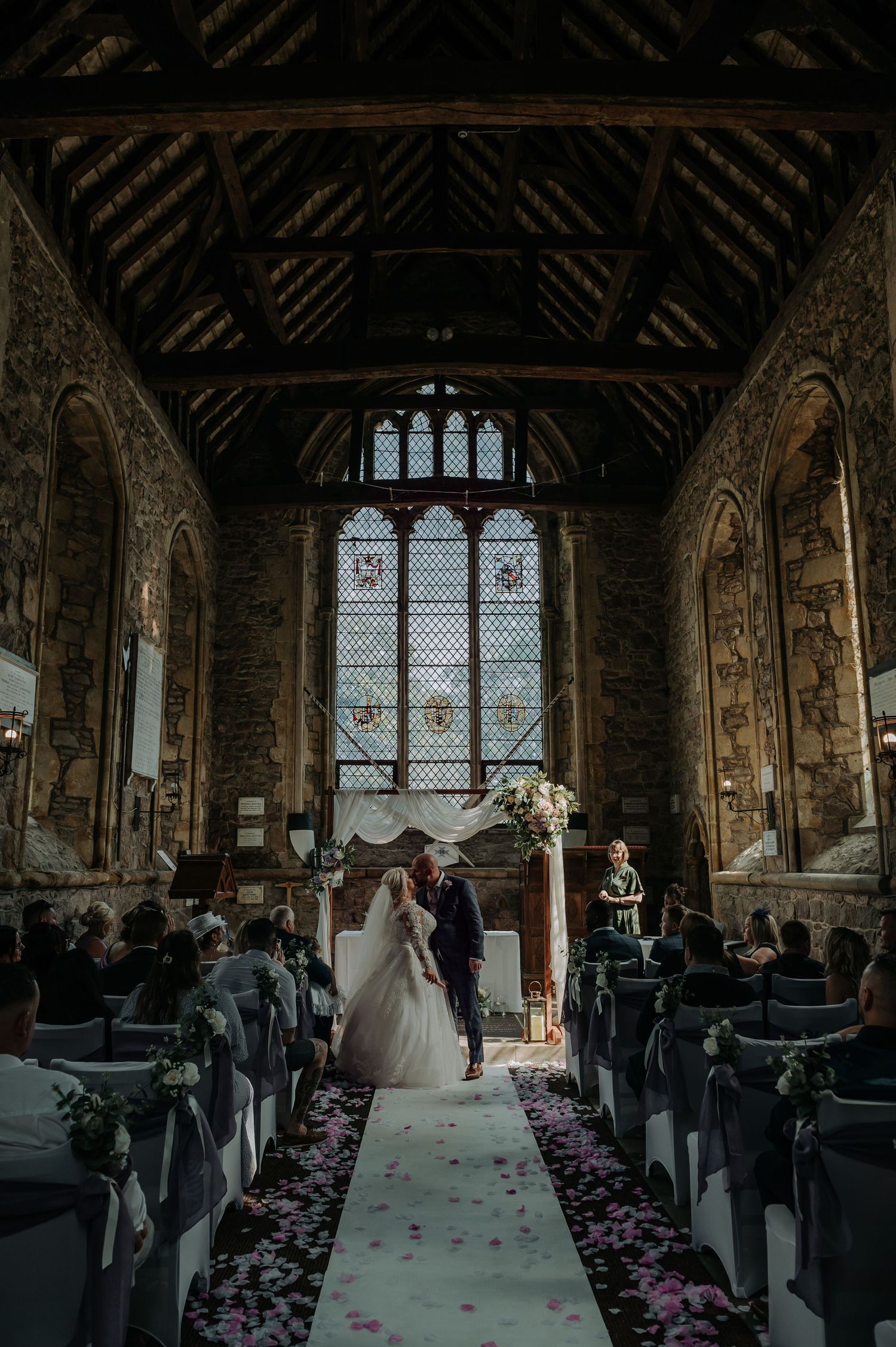 Wedding ceremony taking place in a historic stone church with stained glass windows and rose petals lining the aisle.