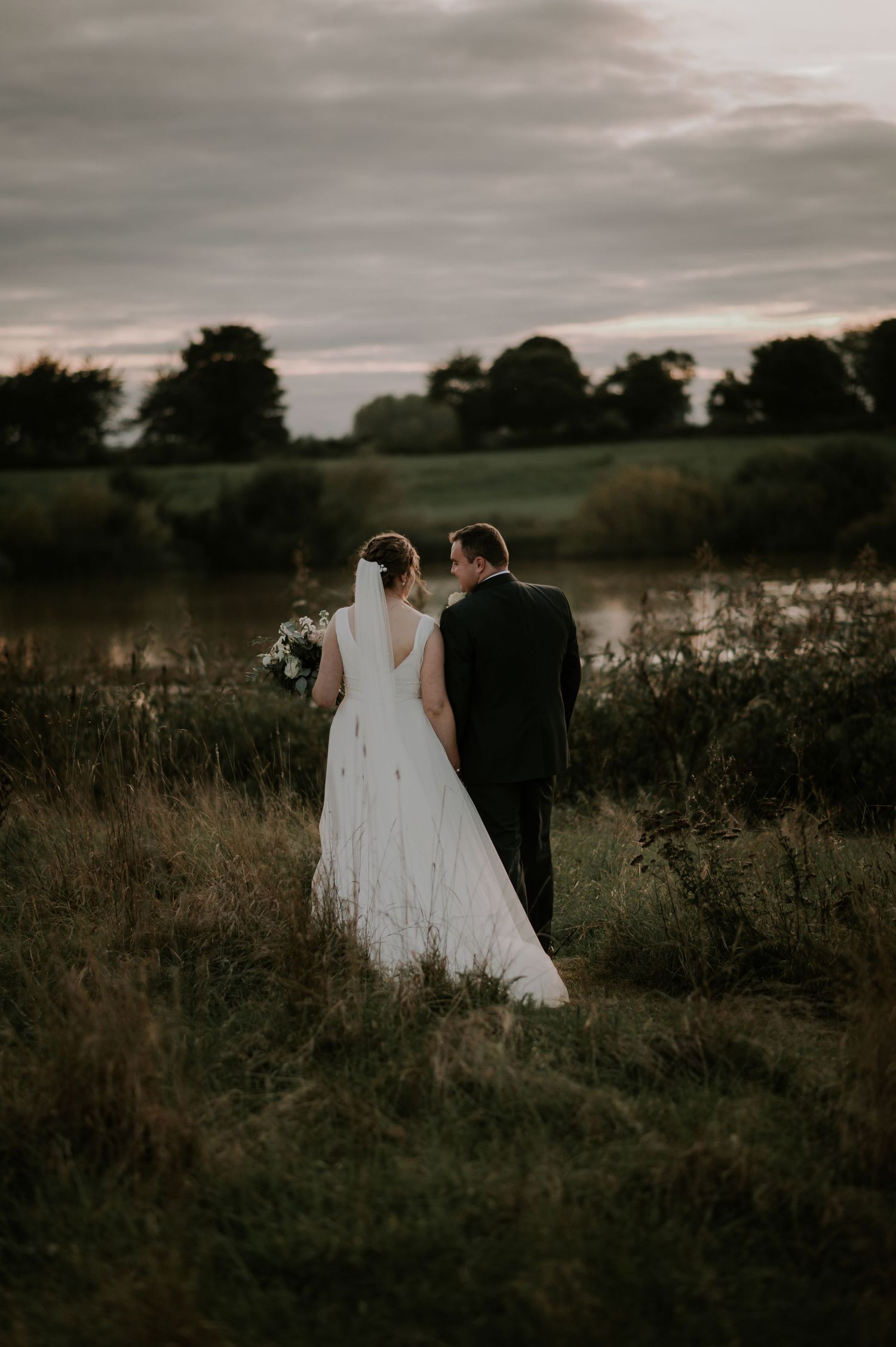 Bride and groom standing together in moody field at sunset with dramatic cloudy sky overhead.