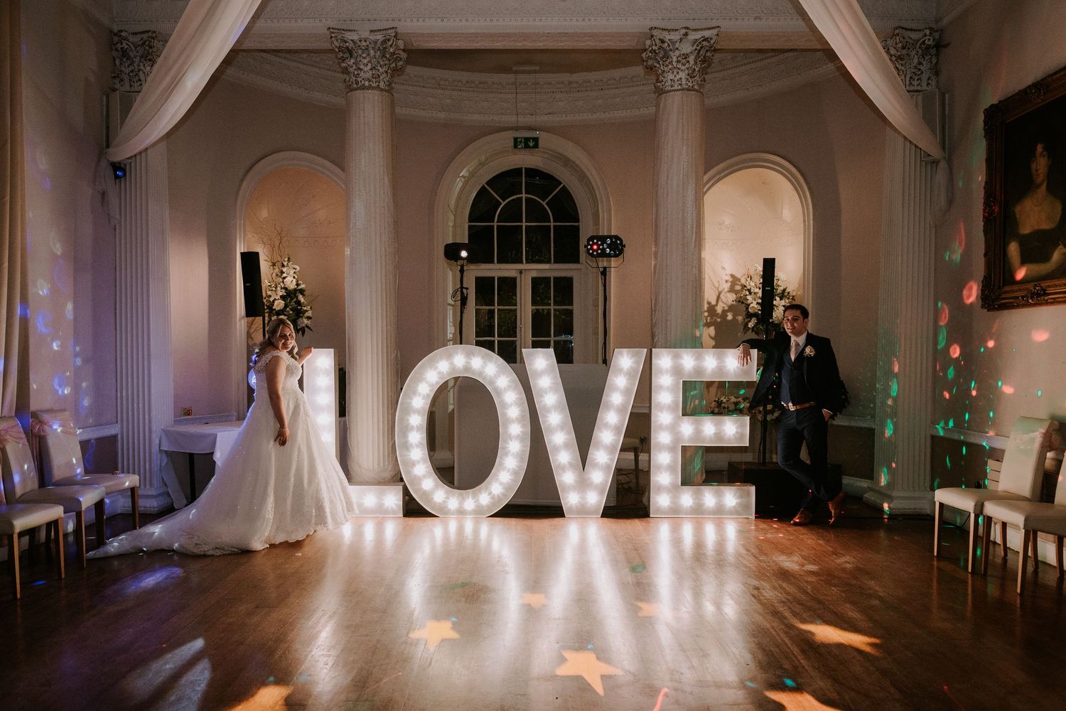 Illuminated LOVE sign glows on dance floor inside elegant room with white columns and arched windows during evening event.