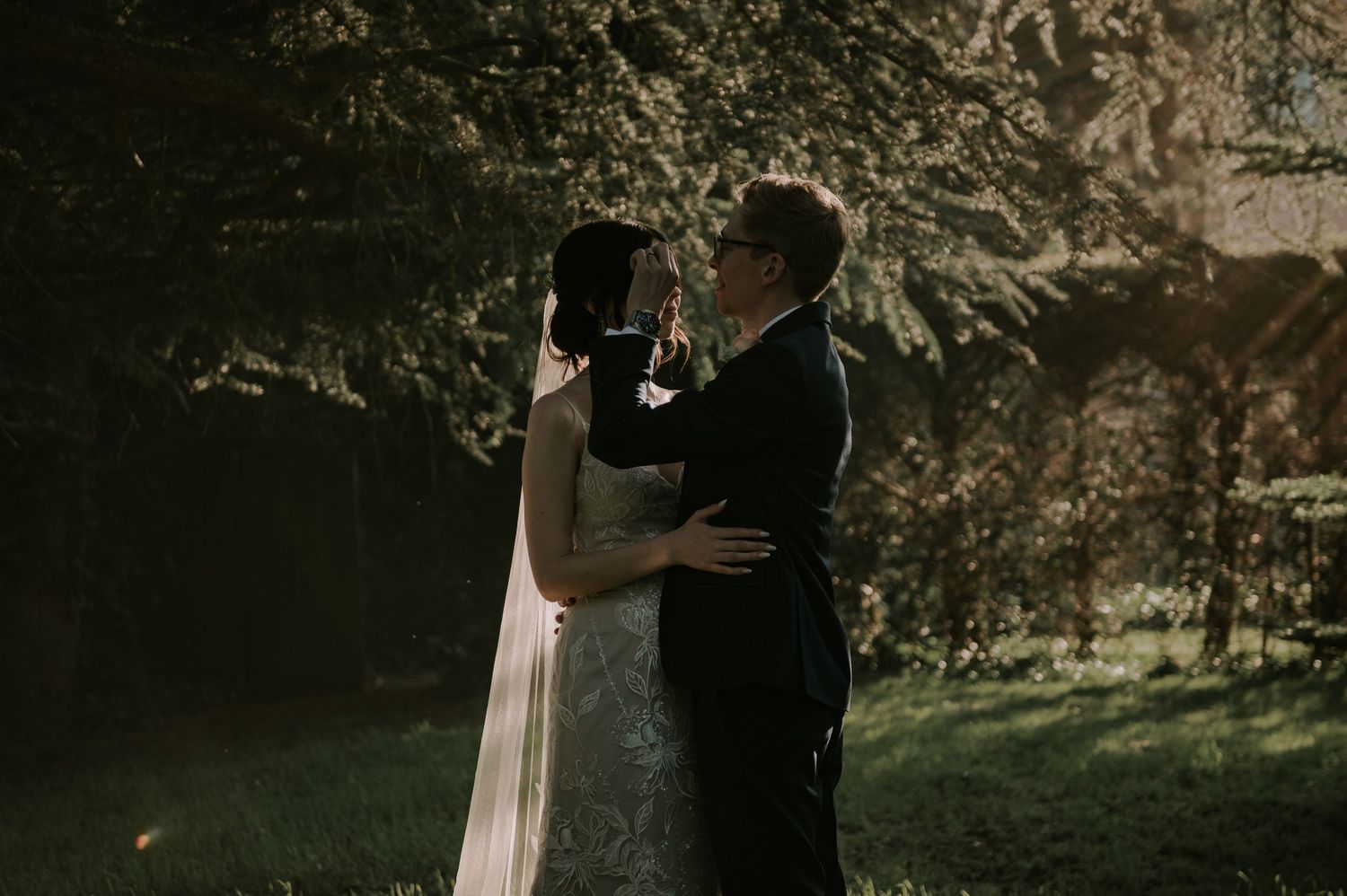 A couple in wedding attire embraces in a romantic moment outdoors with sunlight filtering through trees in the background.