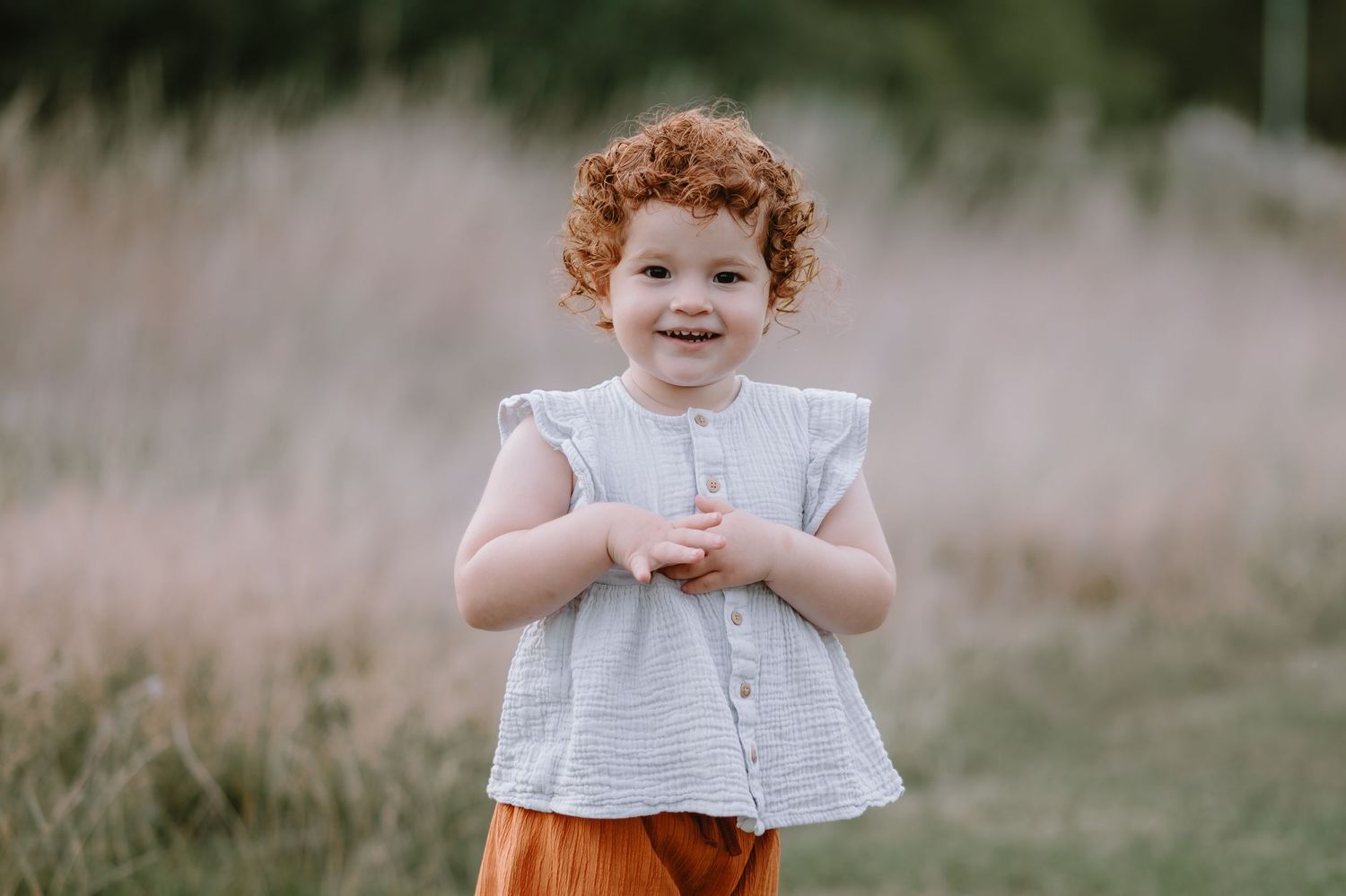 A child with curly red hair wearing a white blouse and orange pants stands in a grassy field on a sunny day.