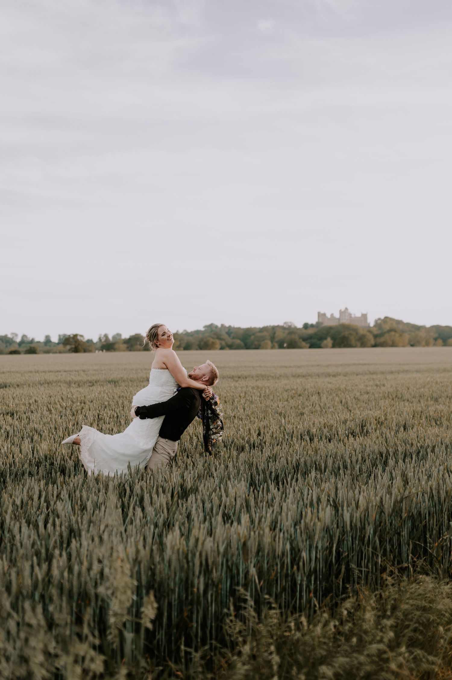 Groom lifting bride in white dress in a lush green wheat field with trees and a castle in the background.