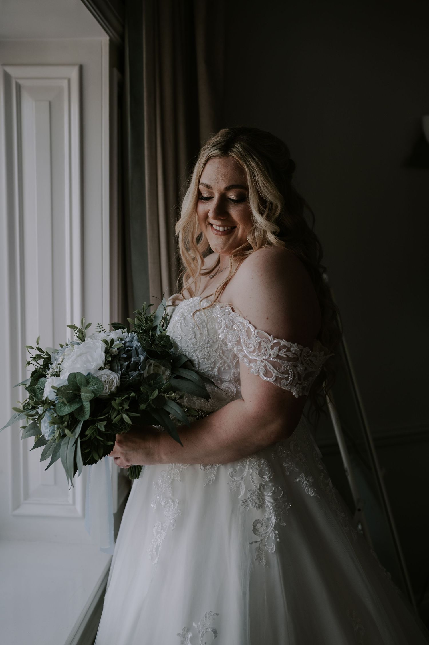 Bride in a lace wedding dress holds a bouquet of eucalyptus and greenery against a moody dark backdrop.