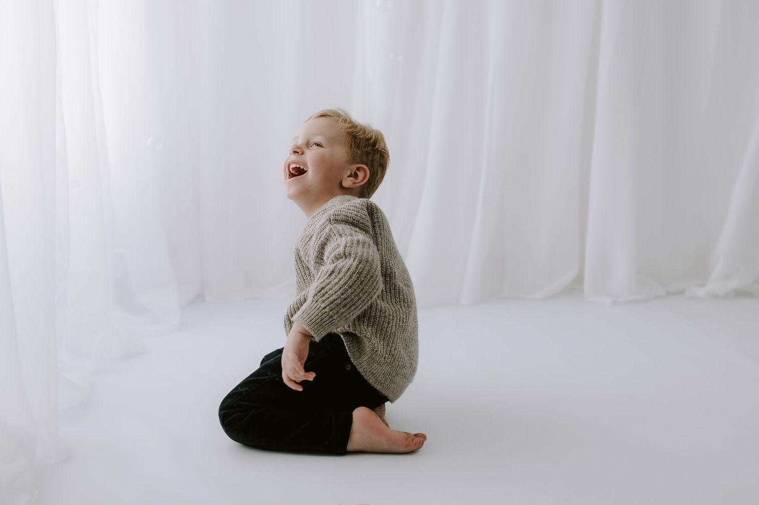 Young child in beige sweater sitting on floor against white curtain backdrop, studio, Melton Mowbray.