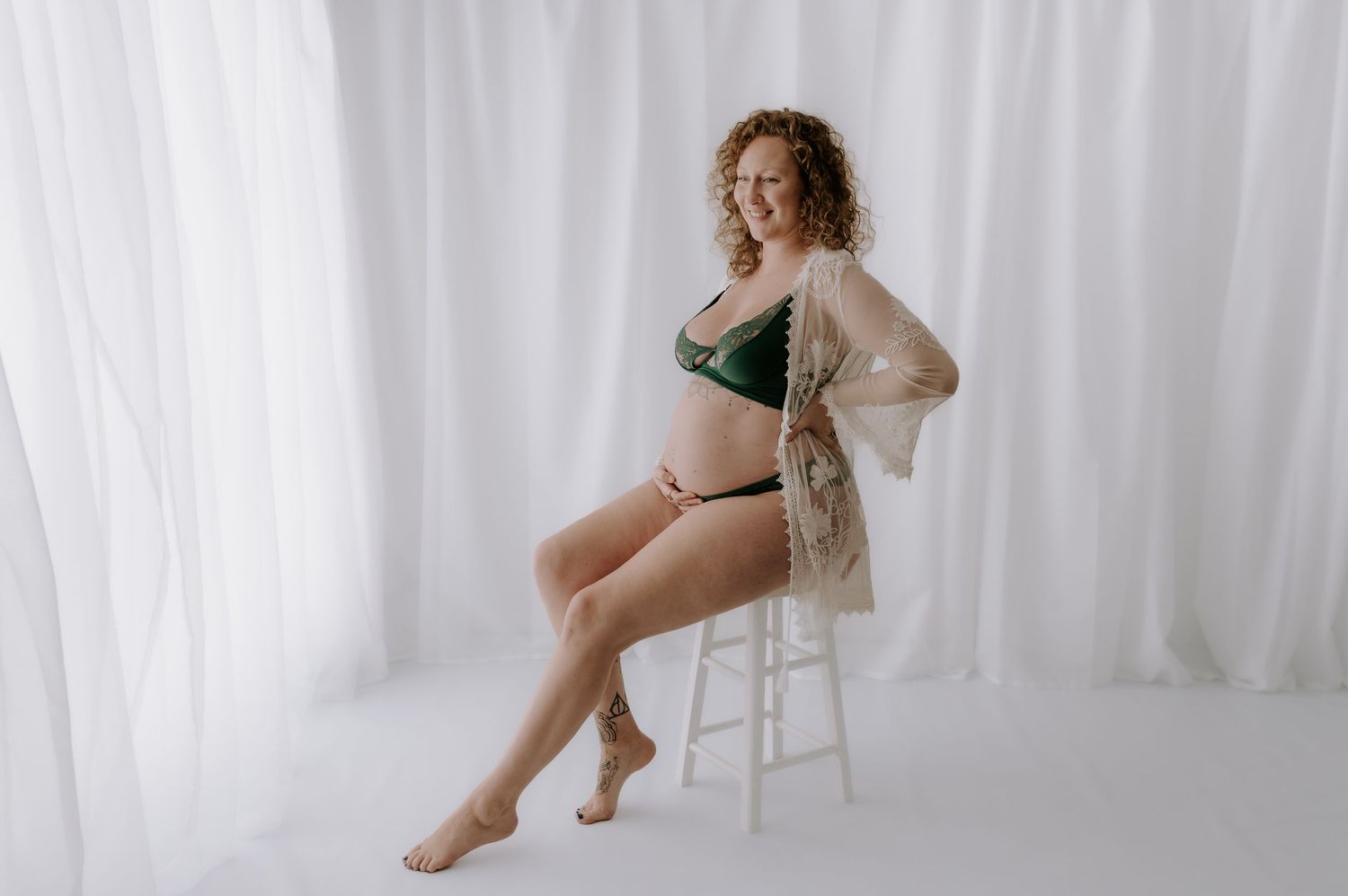 Person in white lace robe and lingerie poses elegantly on white stool against white curtain backdrop.