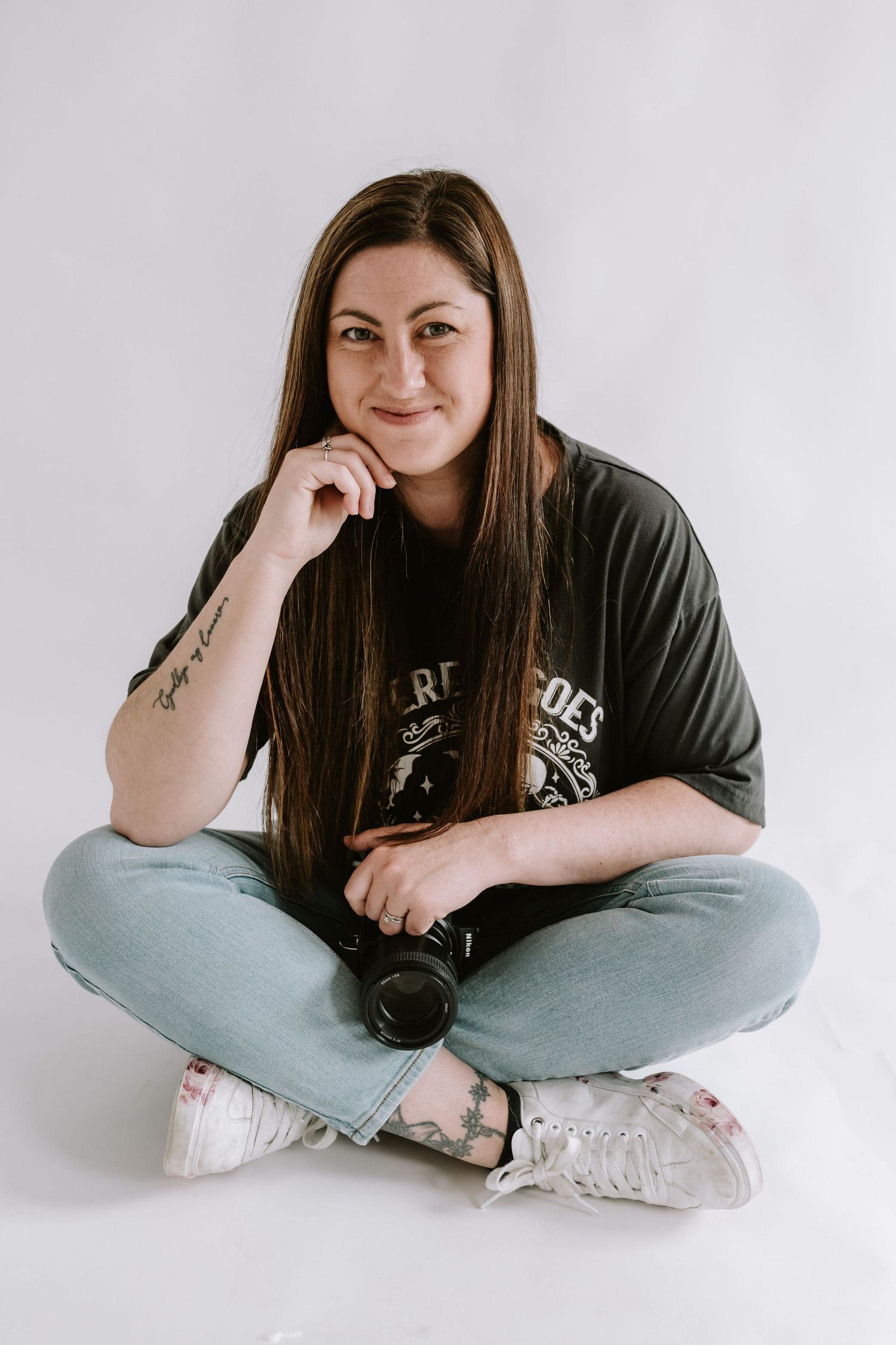 A photographer in a black graphic t-shirt and light jeans sits cross-legged on white backdrop while holding a camera lens.
