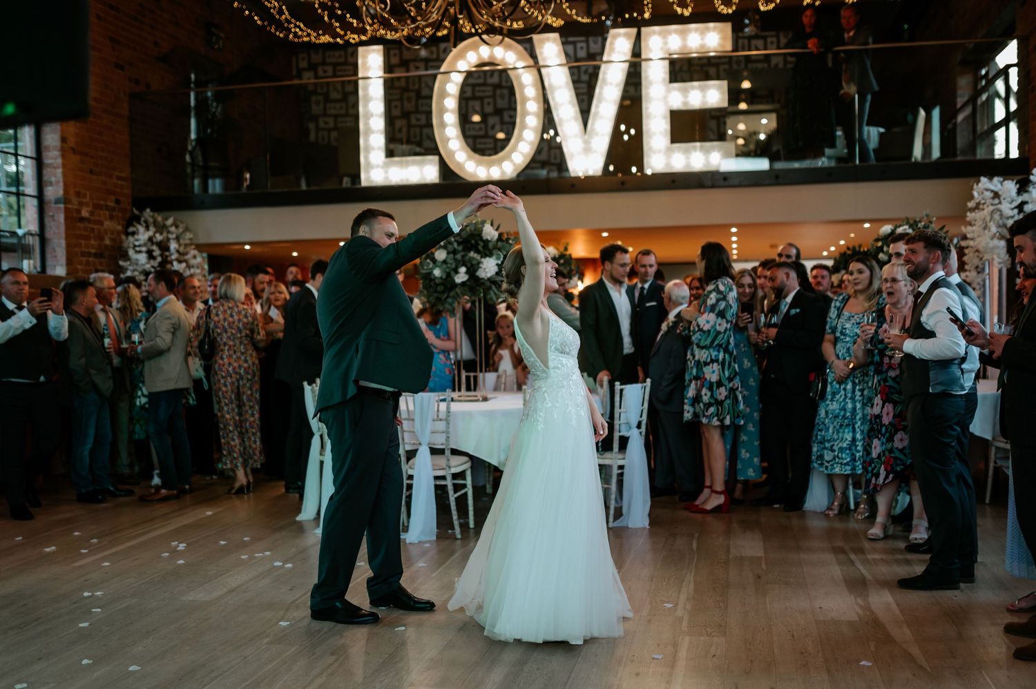 Wedding couple shares their first dance under illuminated LOVE sign as guests watch in a rustic venue with falling confetti.