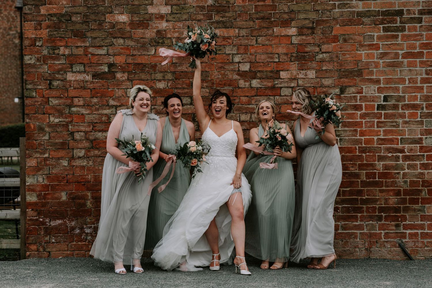 Wedding party in gray bridesmaid dresses sits casually in front of rustic brick wall during outdoor photo session.