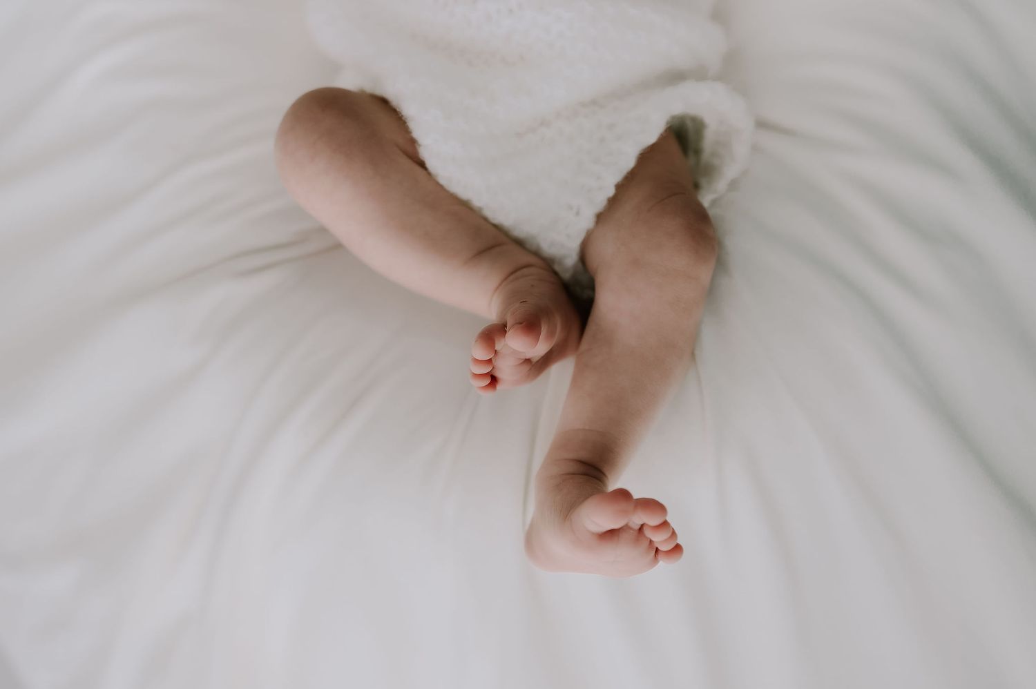 A newborn babies legs kicking the air. Wearing a white romper on a white blanket. Newborn photoshoot in Melton Mowbray