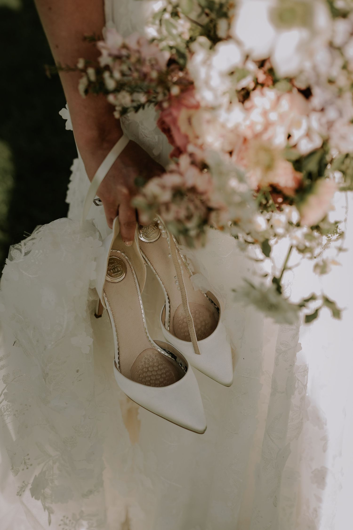 Elegant white bridal shoes held against a flowing wedding dress with soft pink and white floral bouquet in background.