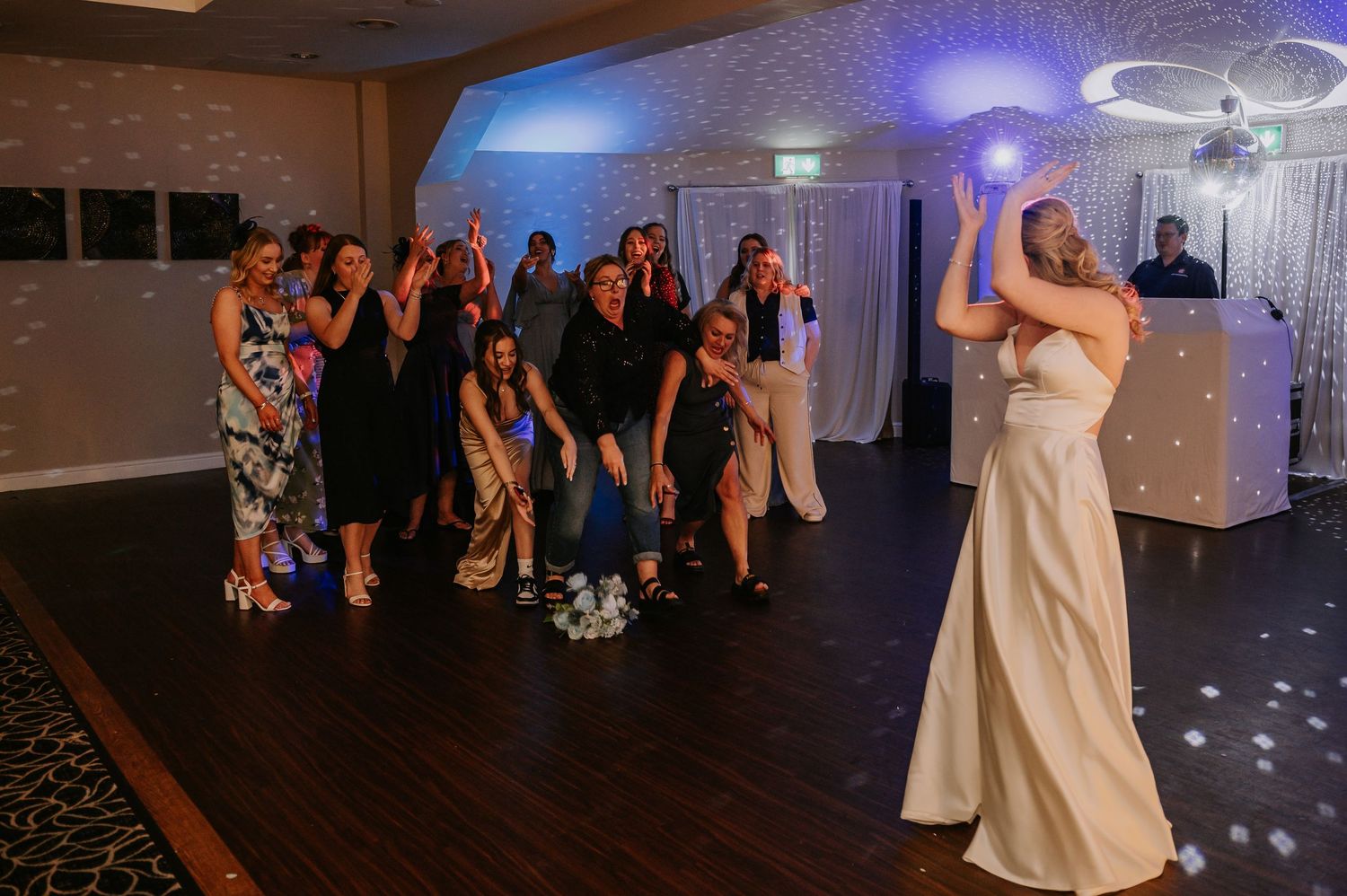 A group gathers at a wedding reception while someone in a white dress throws a bouquet on the dance floor.