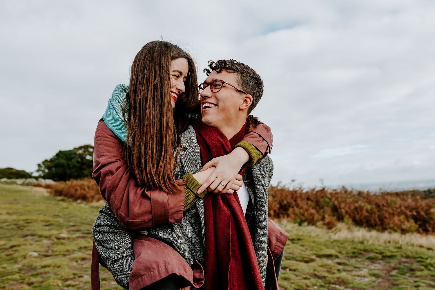 A couple in matching burgundy jackets embrace and laugh together while standing in an autumn field under cloudy skies.