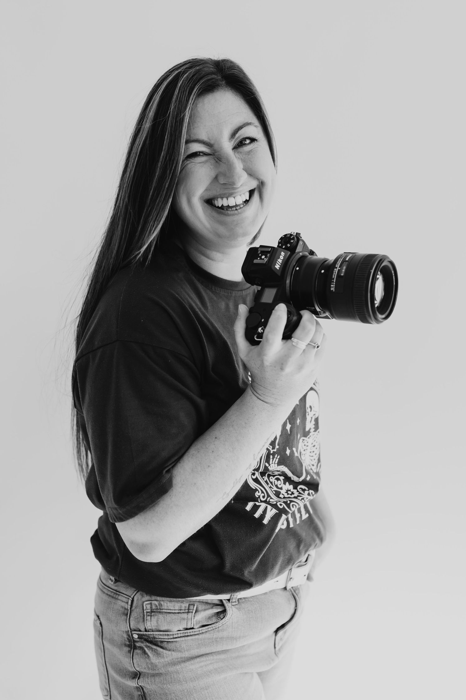 A photographer in denim overalls and black shirt laughs joyfully while holding a camera in black and white.