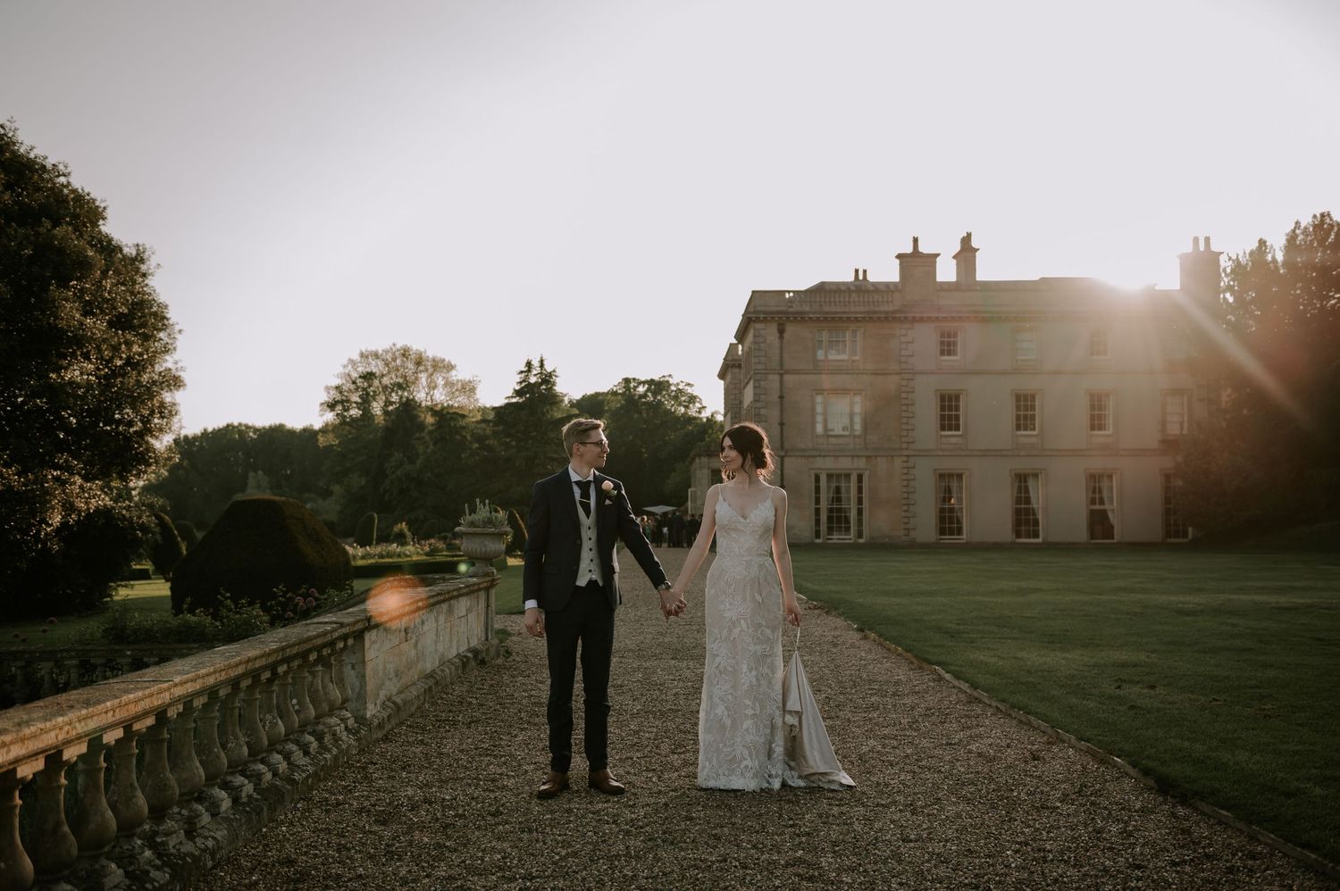 A couple celebrates their wedding day in front of an elegant stone mansion at sunset on a manicured lawn.