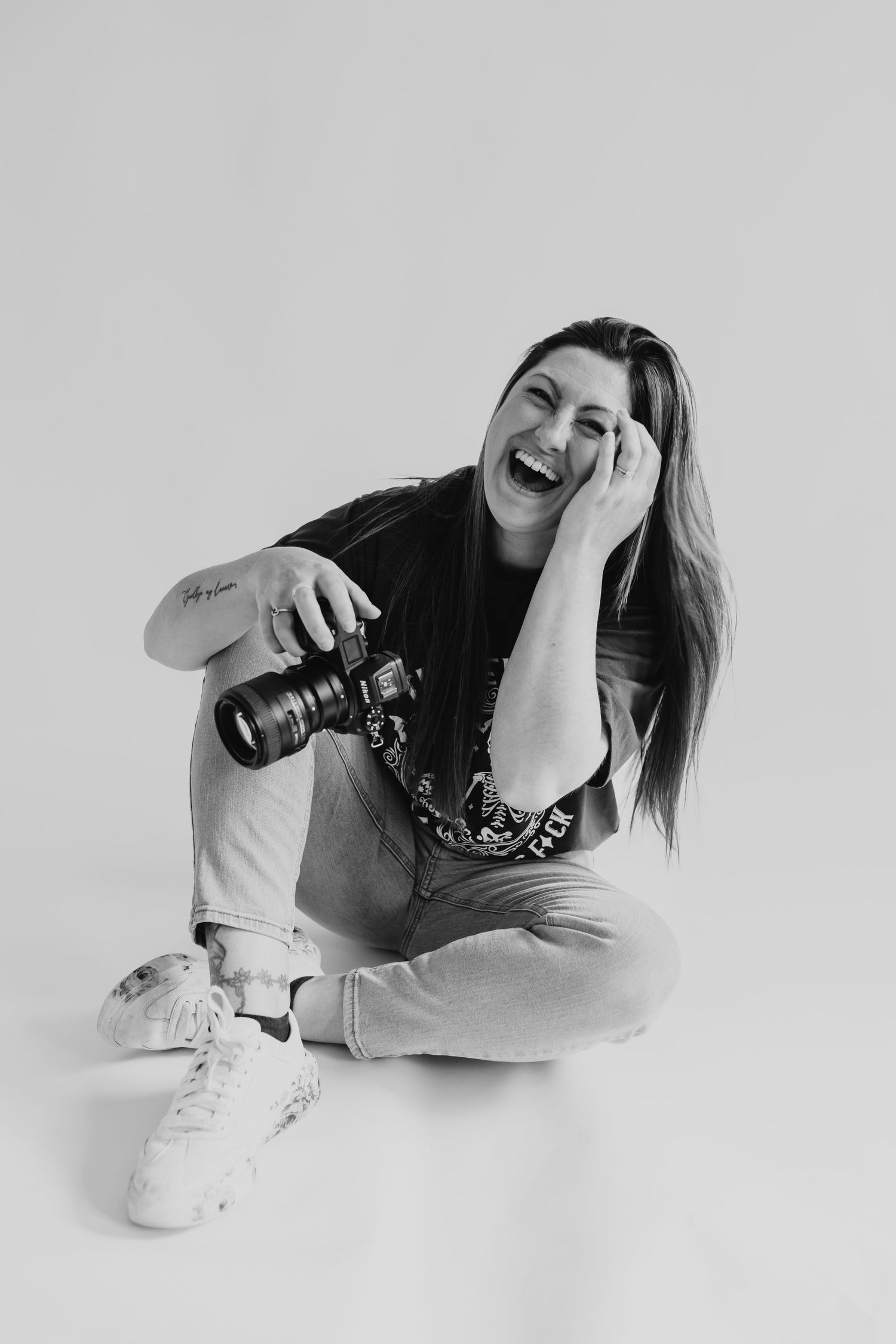 Black and white photo of photographer with camera laughing while sitting cross-legged against white background.