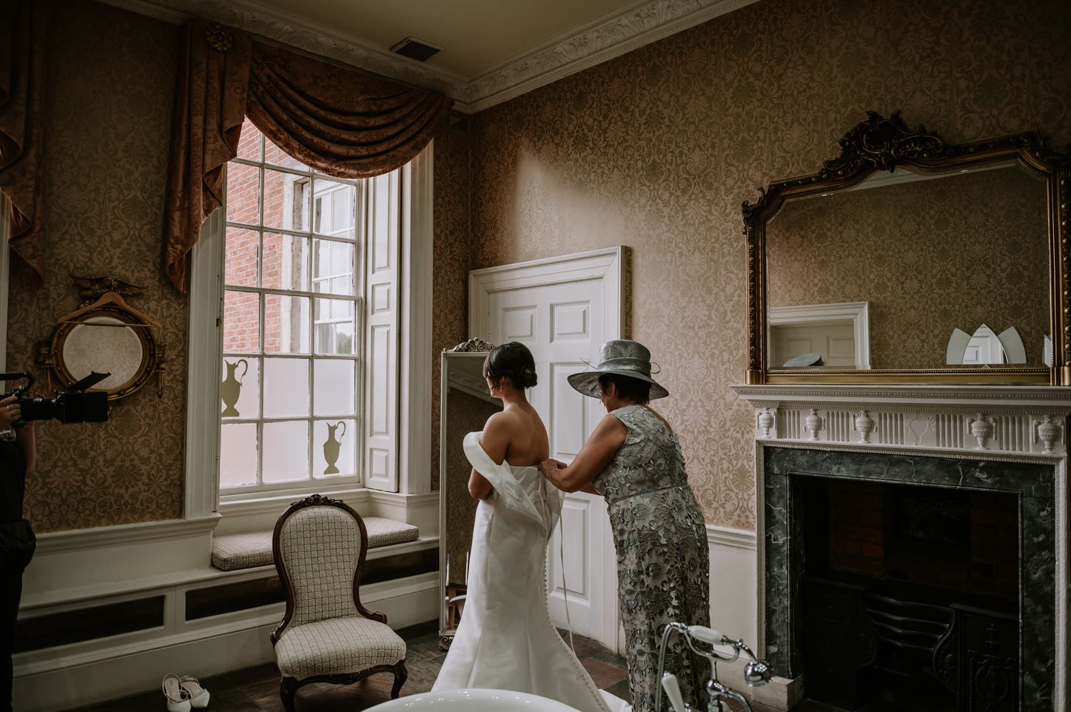Two people stand by an elegant window in a vintage room with ornate mirrors and a fireplace during a special occasion.