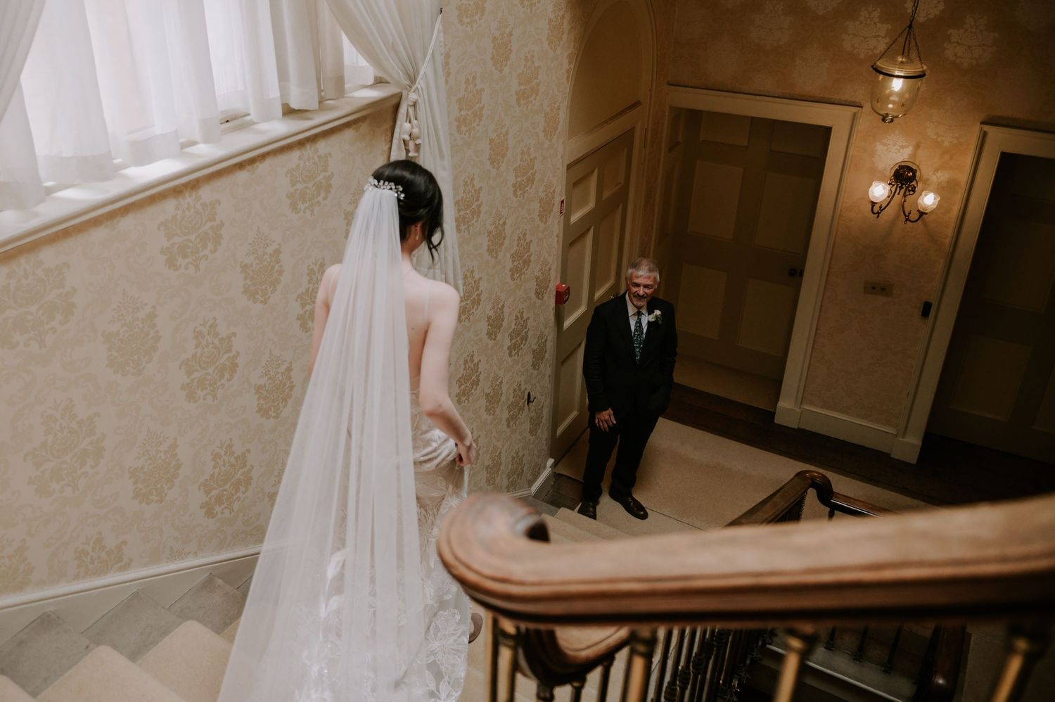 A wedding couple descends an elegant curved staircase in a grand venue adorned with crystal chandeliers.