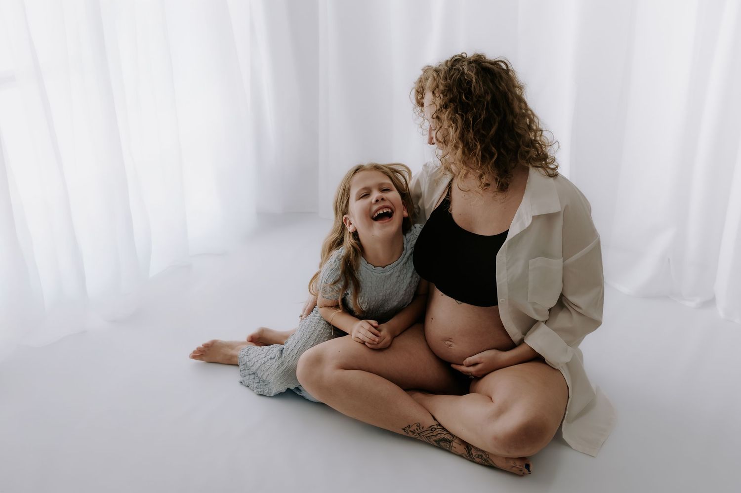 Little girl laughing with pregnant mum on white floor, both smiling, studio, Melton Mowbray.