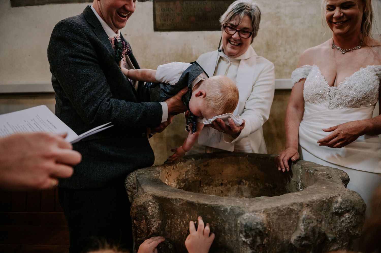 Baby being baptized in a stone font during a wedding ceremony with family members gathered around.