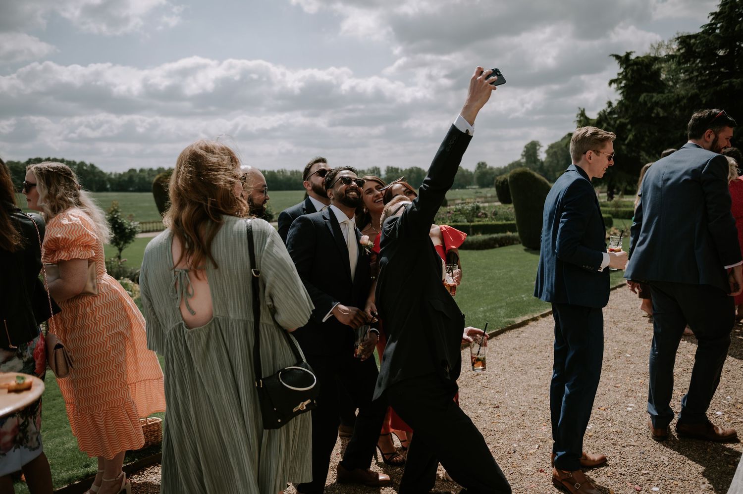 A group of wedding guests gather outdoors on a grassy lawn under cloudy skies during a summer celebration.