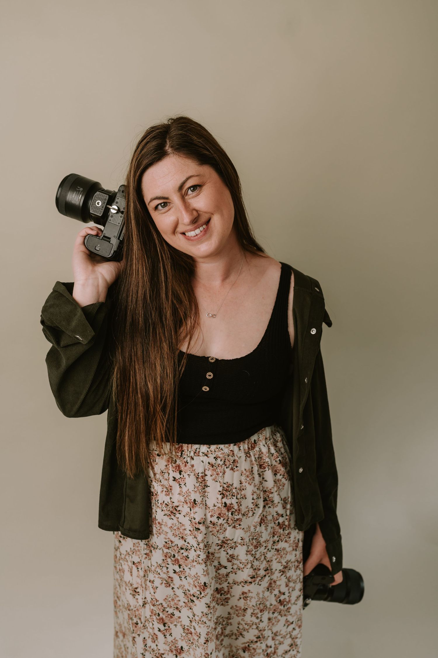 Professional photographer in olive jacket holding camera and lens against beige wall while wearing floral skirt.