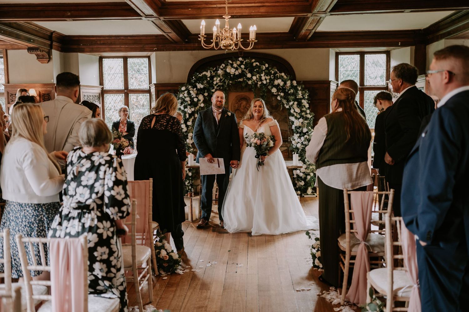 Indoor wedding ceremony taking place under floral arch in rustic venue with exposed wooden beams and natural lighting.