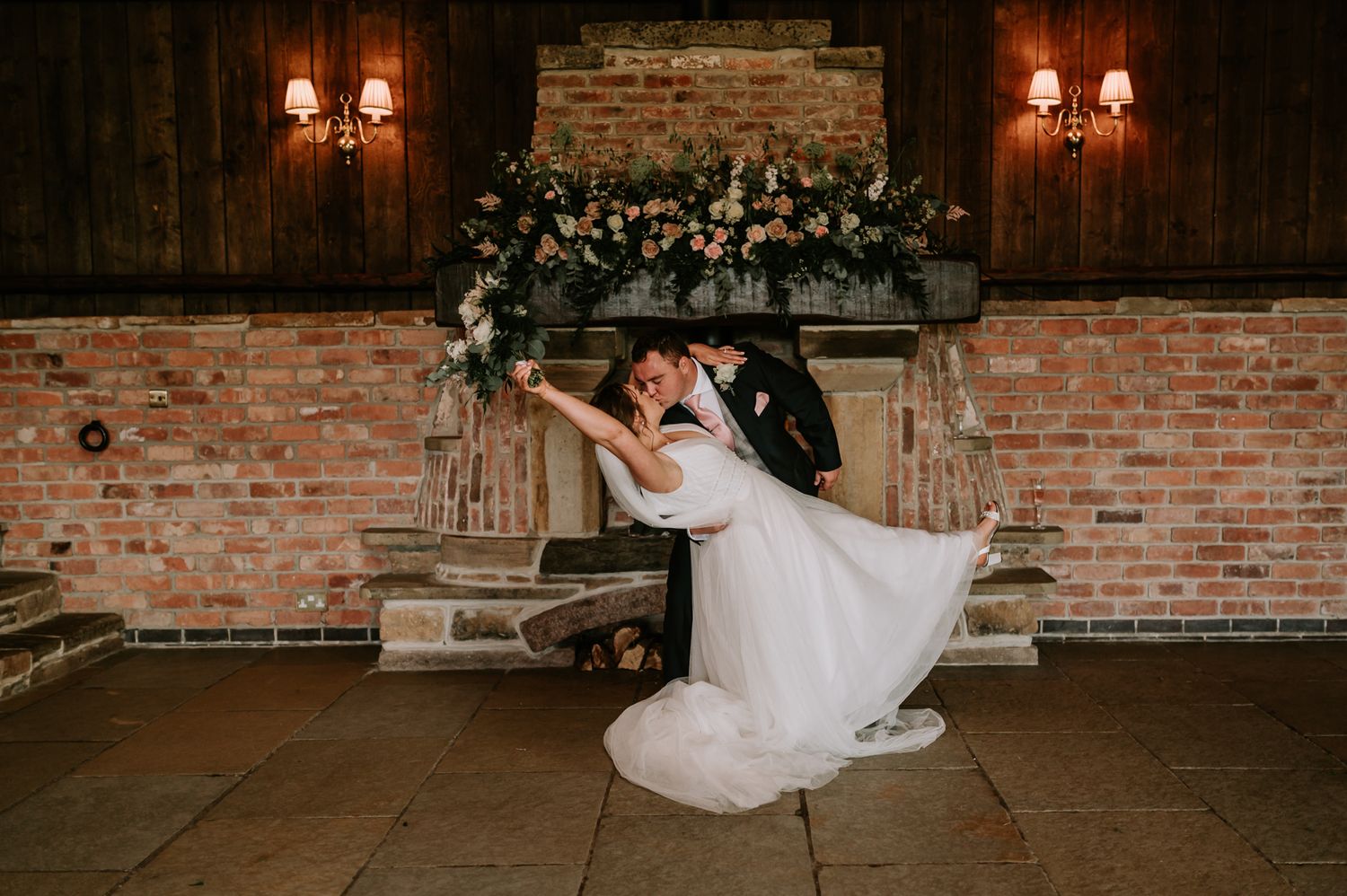 A romantic dance pose in a rustic brick-walled venue with warm lighting and floral decorations.