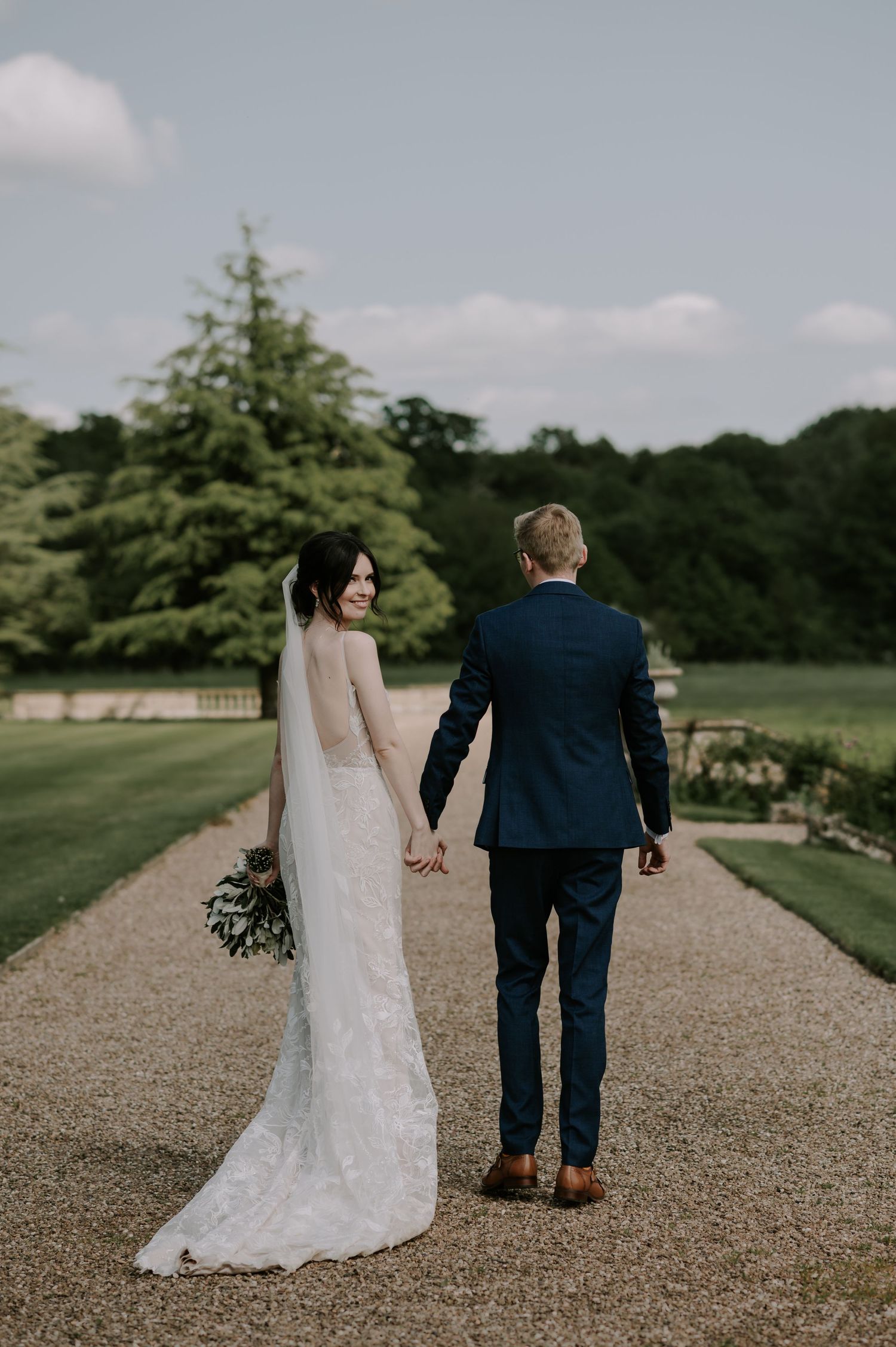 Newlywed couple walking together on a gravel path through scenic countryside gardens.