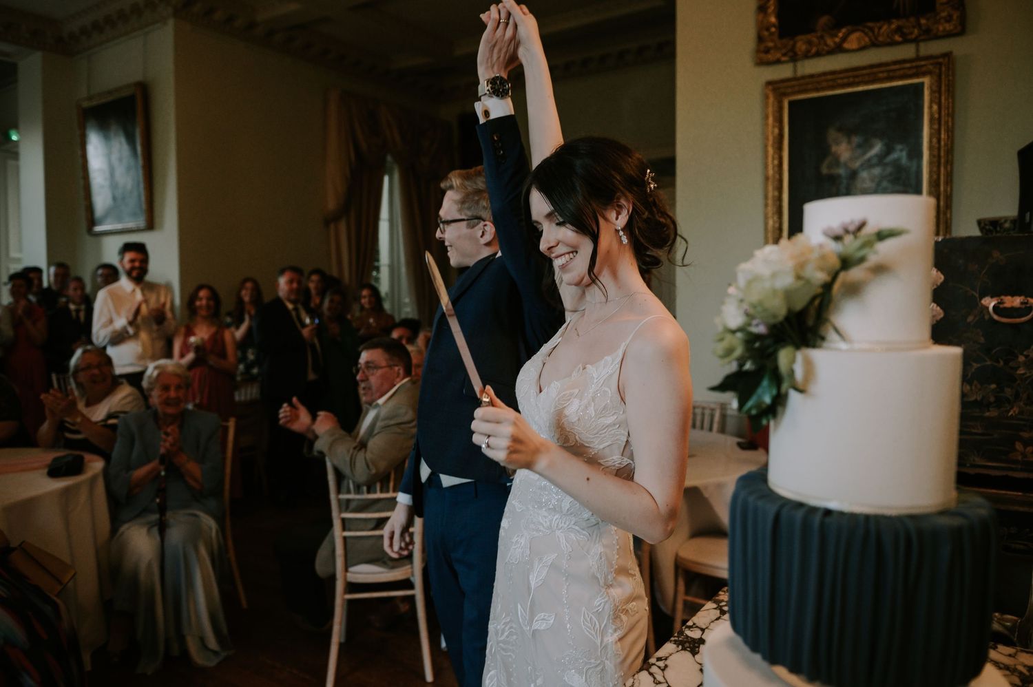 Newlyweds celebrate with a toast and raised hands during their wedding reception next to an elegant white tiered cake.