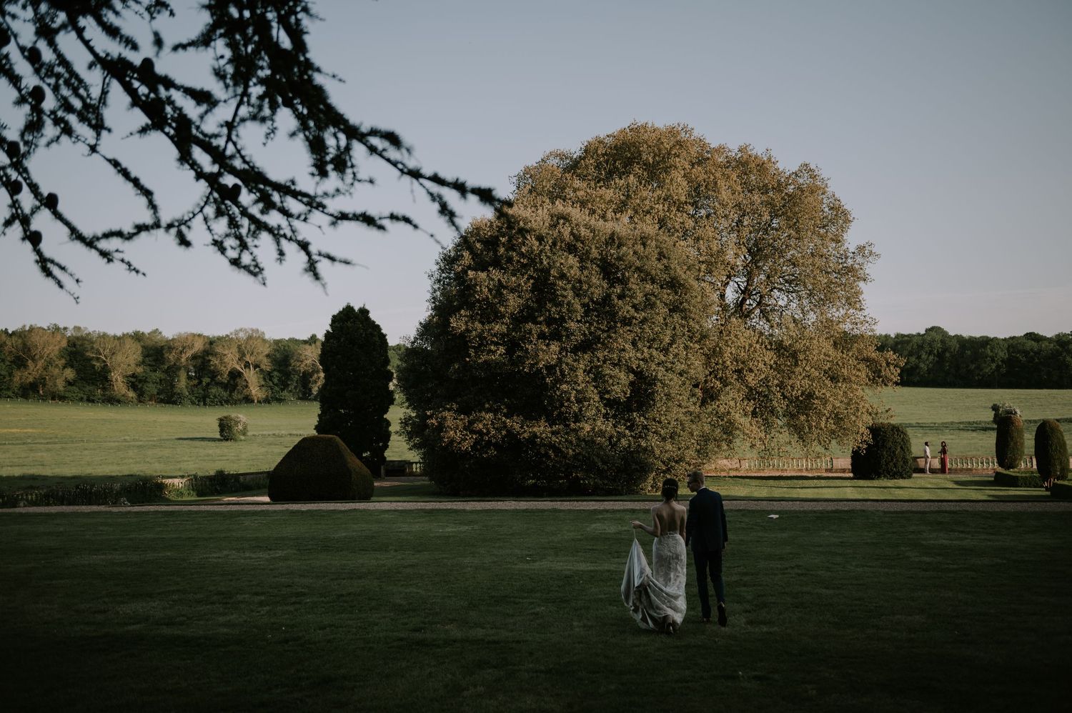 Wedding ceremony attendees walk across a green field with autumn trees in the background at sunset.