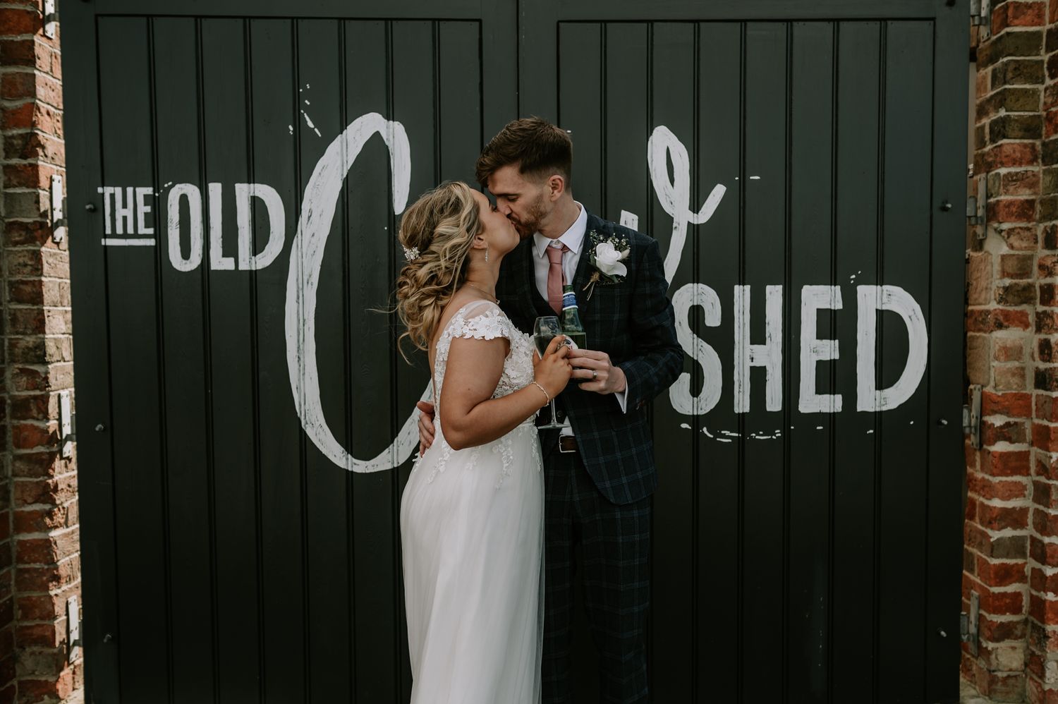 A couple in wedding attire embraces in front of a dark green door with 'The Old Shed' text at a rustic brick venue.