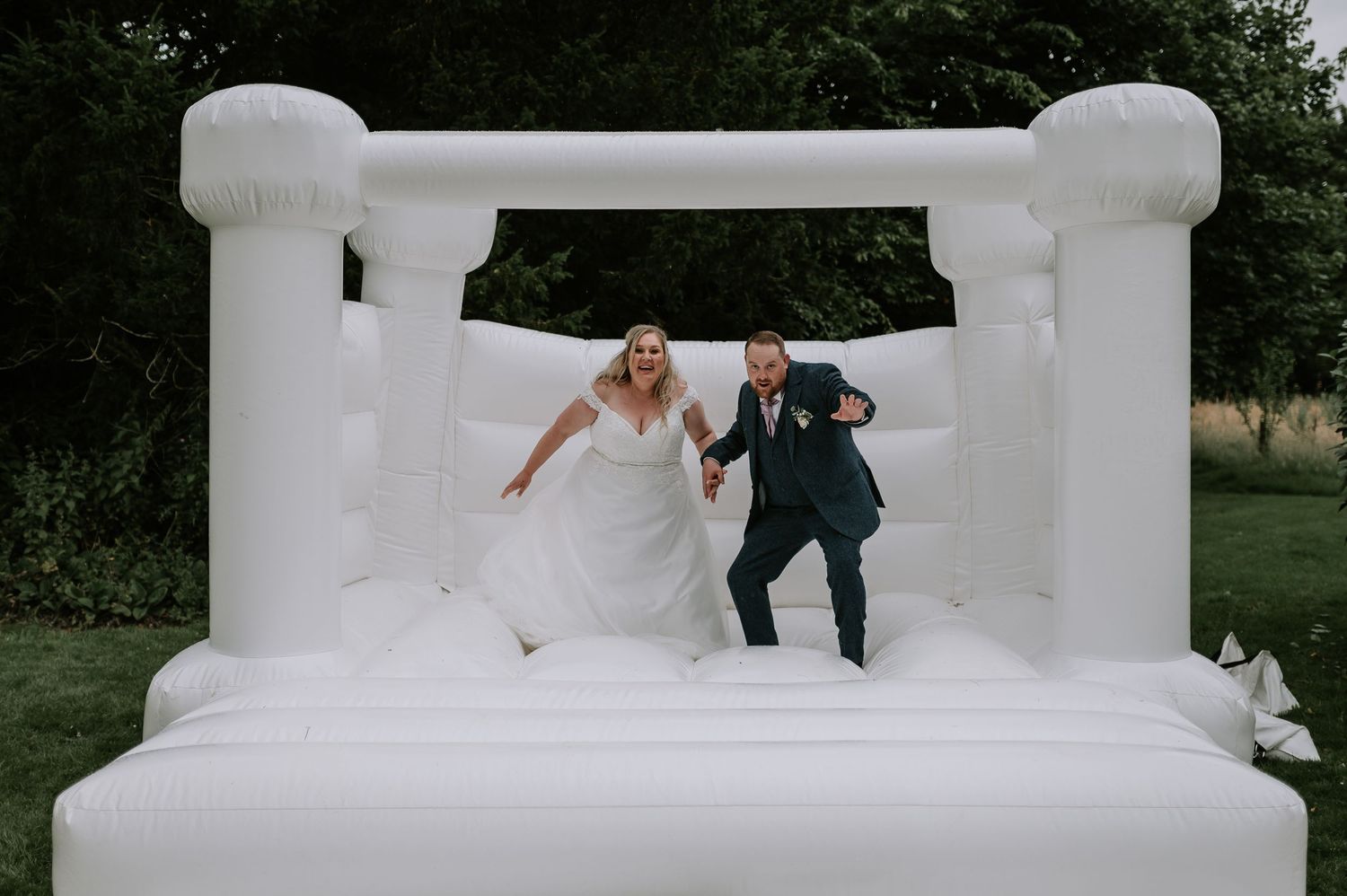 Two people in formal attire joyfully bounce inside a large white inflatable bounce house in an outdoor setting.