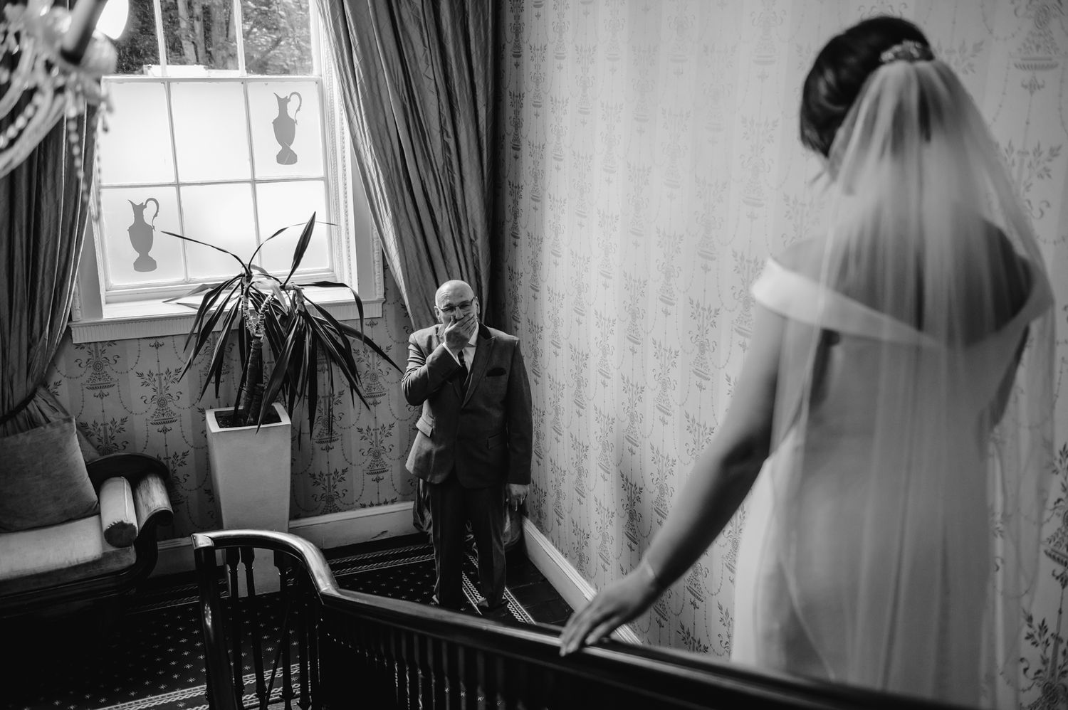A black and white wedding photo showing a tender moment between two people on an elegant staircase with window light.