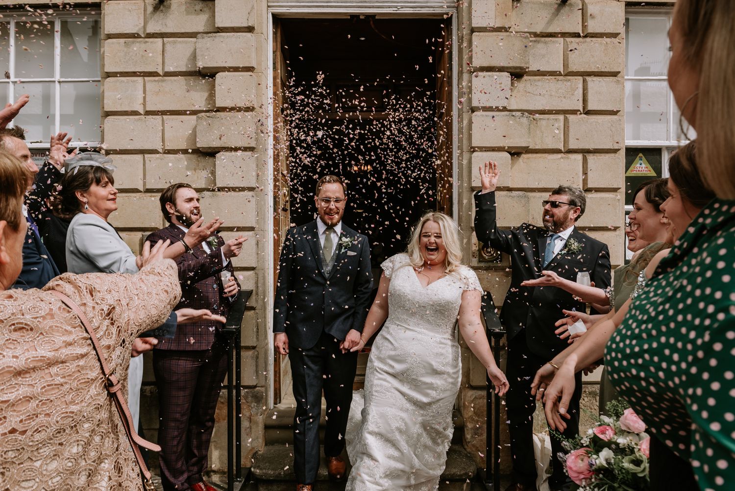 Wedding couple exits venue through doorway while guests shower them with confetti at a historic stone building.
