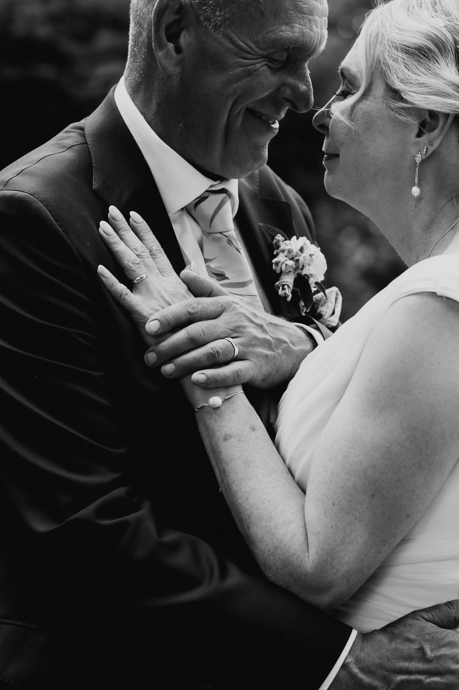 Black and white photo of an older couple in wedding attire smiling and embracing, showcasing their wedding rings.