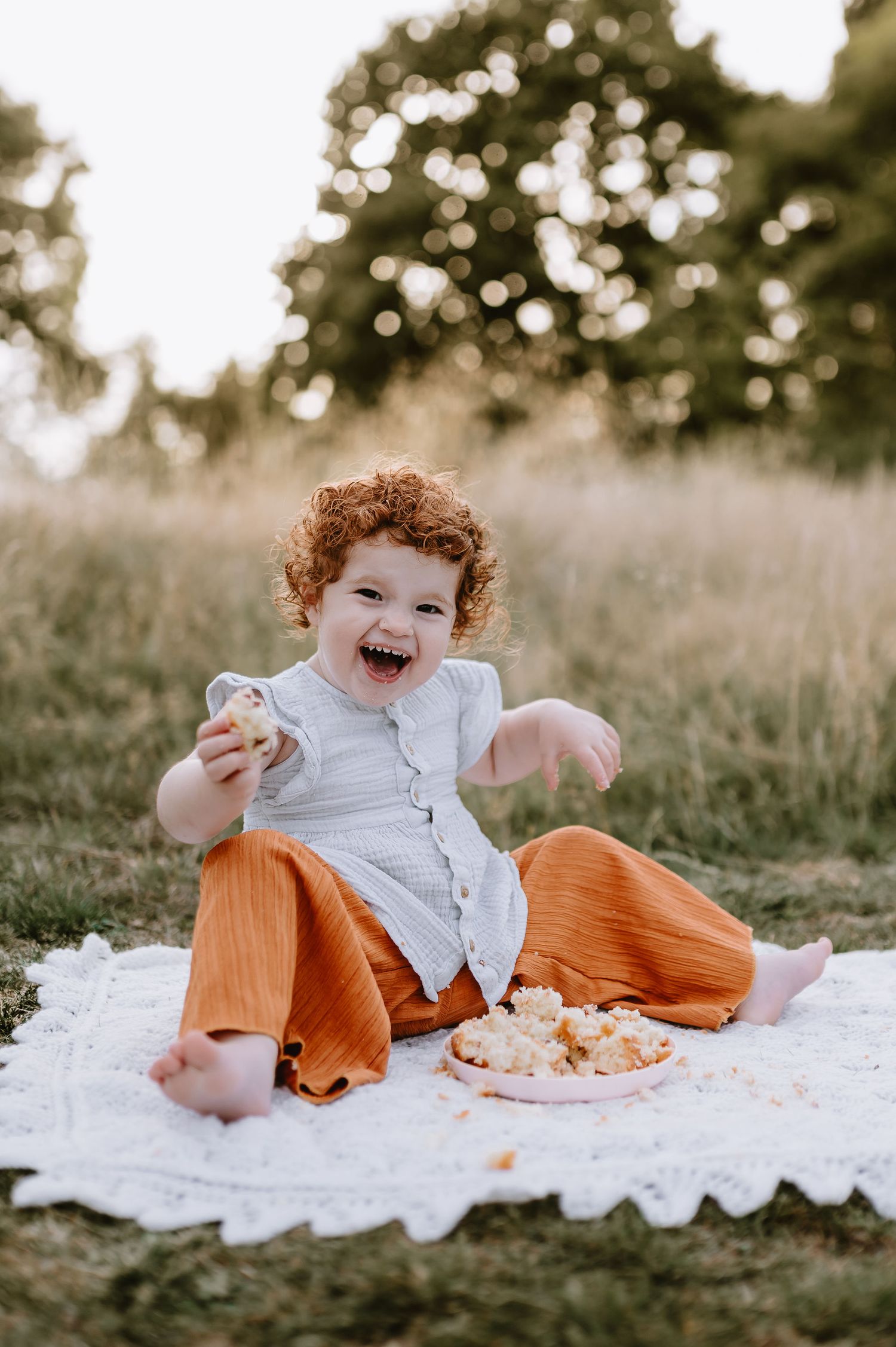Child in orange pants lying on a white blanket outdoors, laughing and holding cake in a grassy field during golden hour.