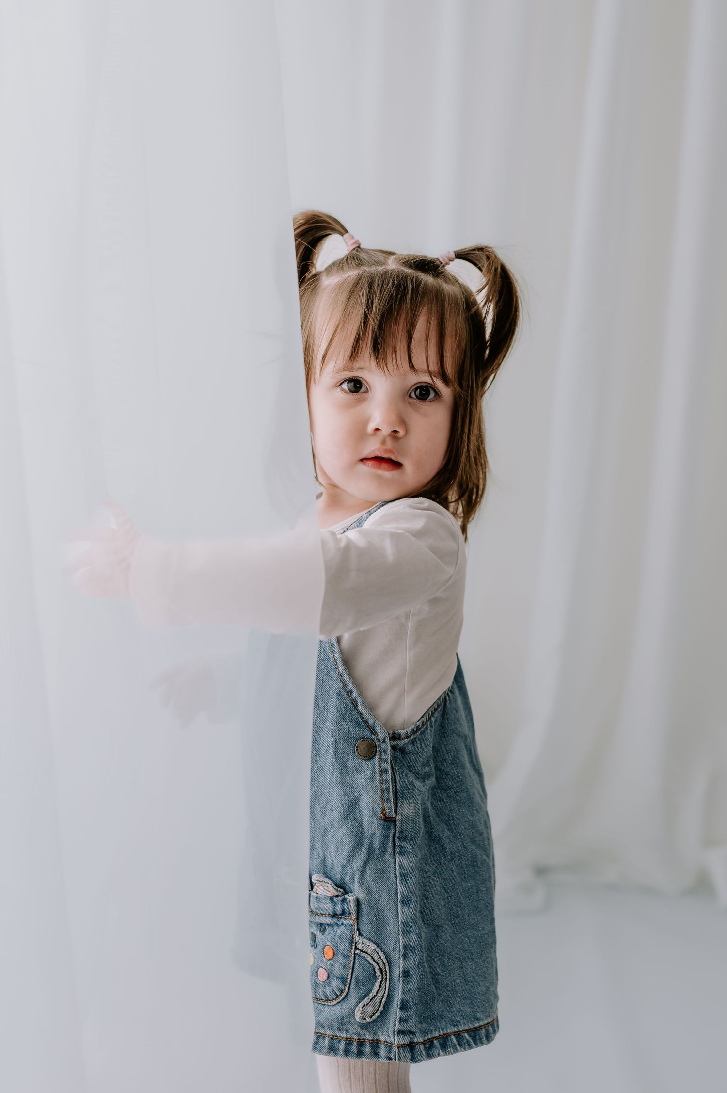 A small child in a denim vest and light top stands against a white curtain background in three similar poses.