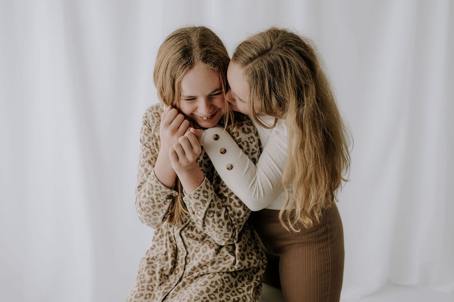 Two sisters laughing together on white stool in bright studio – Melton Mowbray children’s photoshoot.