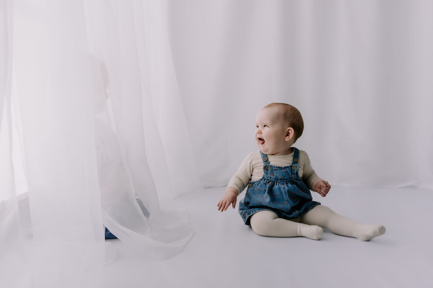 Small child sitting in a white studio, smiling at a young boy hiding behind a curtain. Children's photoshoot Melton Mowbray
