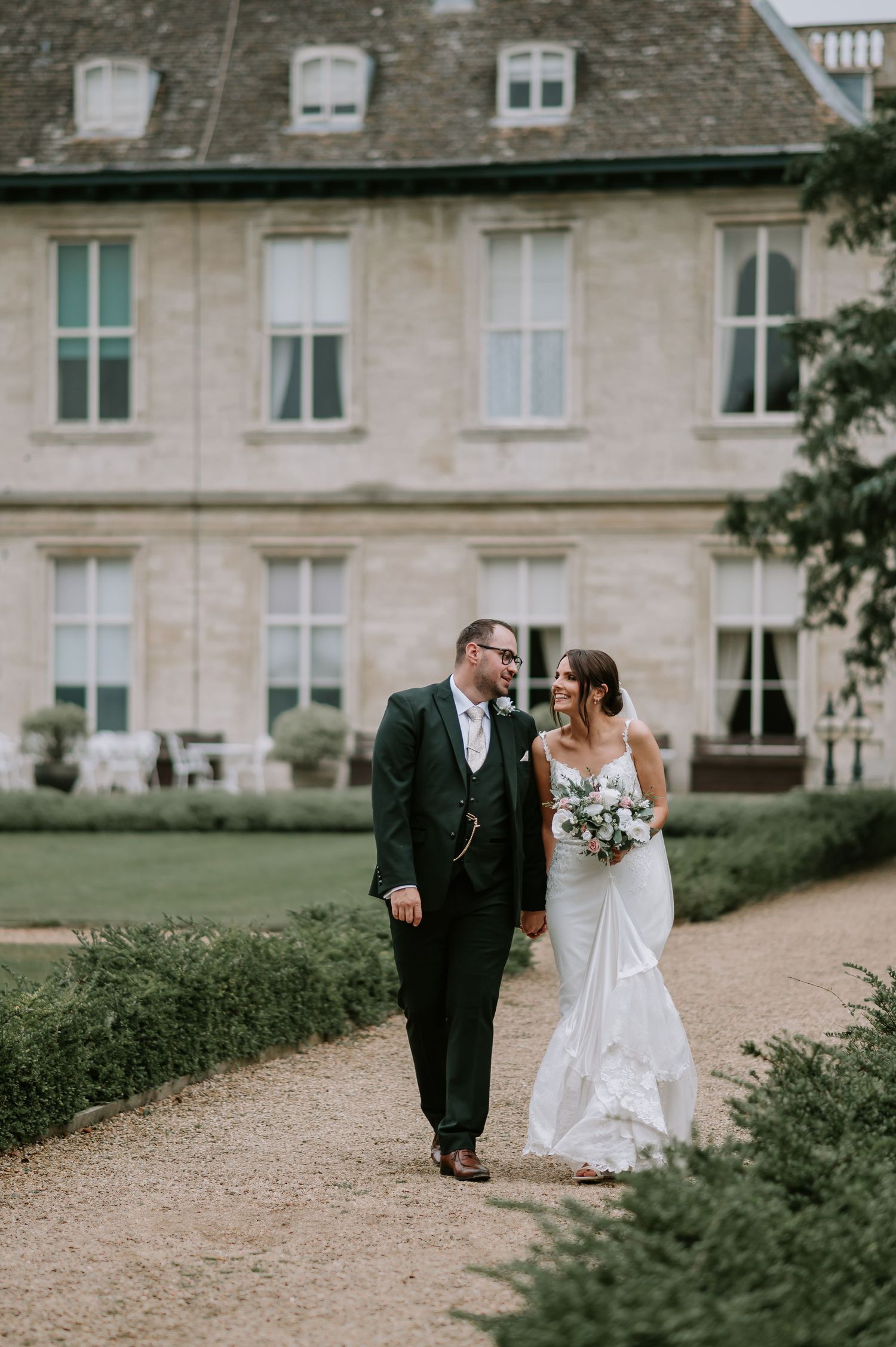 A couple on their wedding day walking along a gravel path outside an elegant stone mansion with dormer windows.