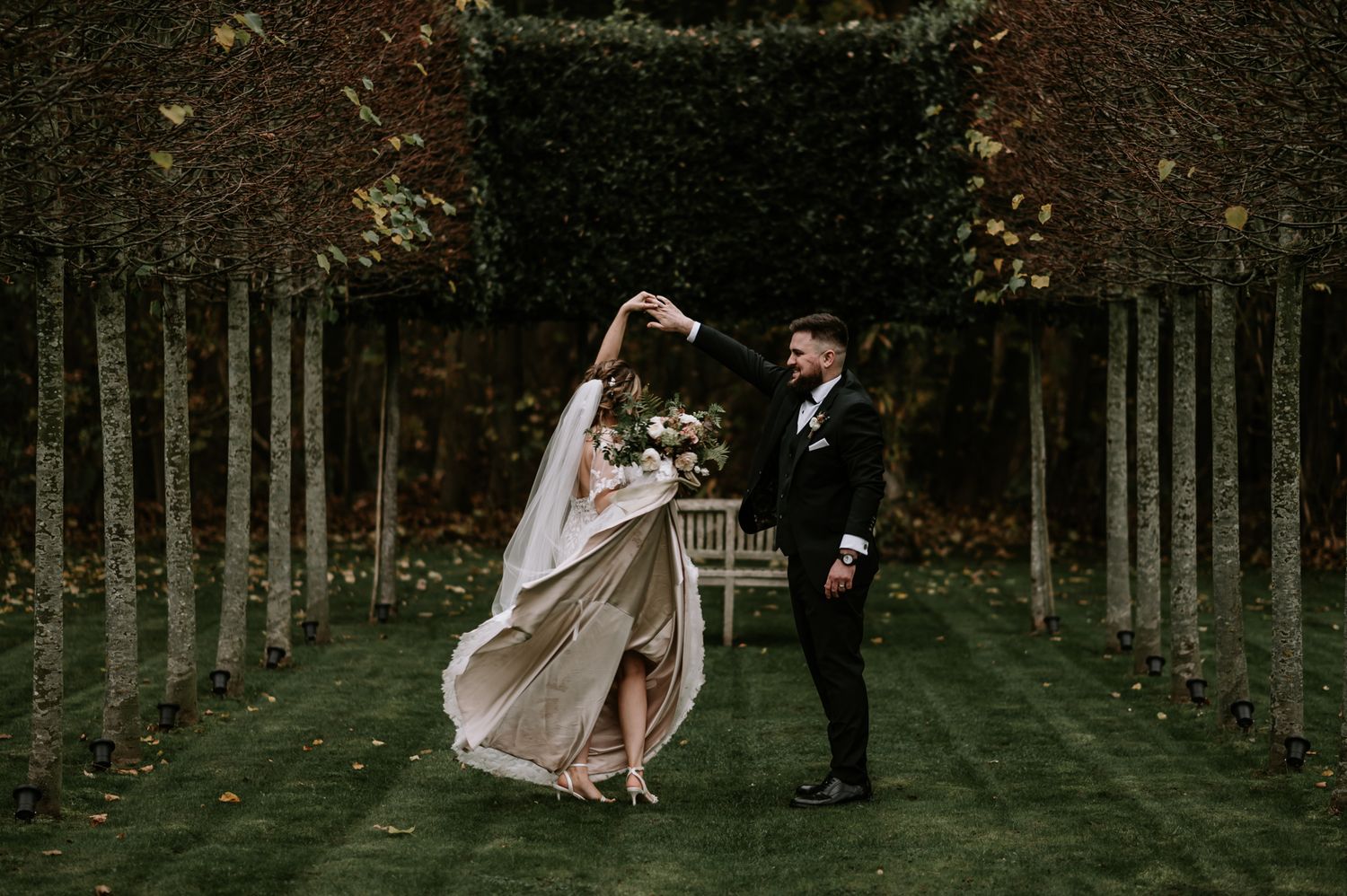 A romantic wedding couple dance together in a lush garden path lined with tall green hedges and trees.