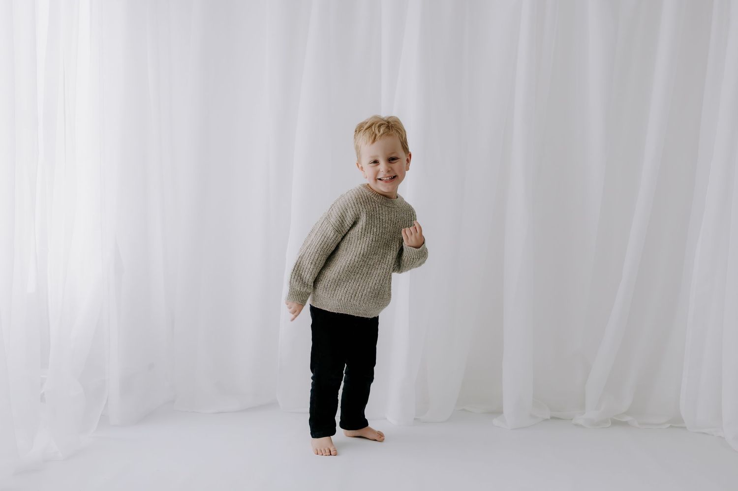Toddler in beige sweater and dark pants standing smiling against white curtain backdrop, studio, Melton Mowbray.