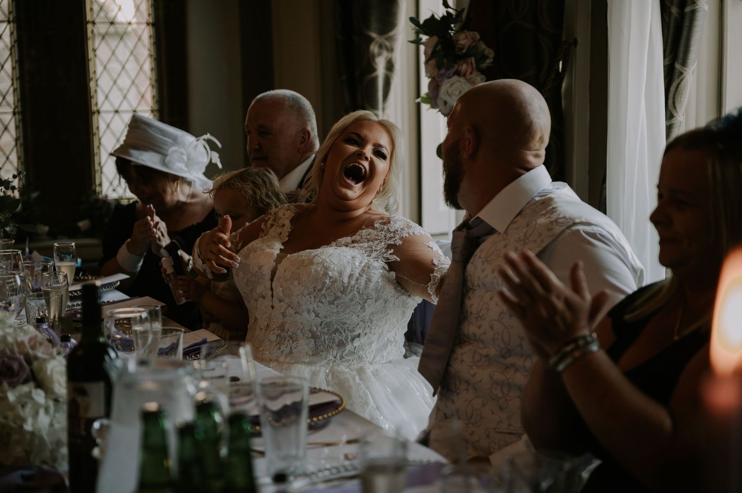 Bride laughing loudly during wedding speeches in sunlit venue, Melton Mowbray.