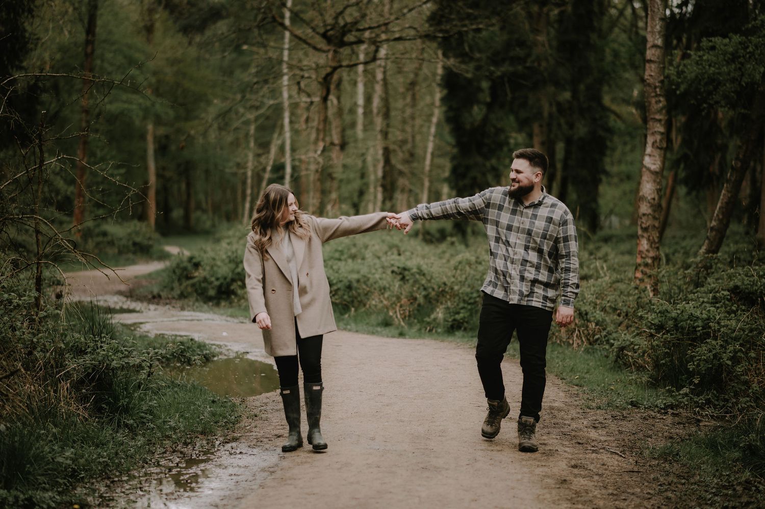 A couple holds hands while walking down a forested path in matching plaid and casual outfits.
