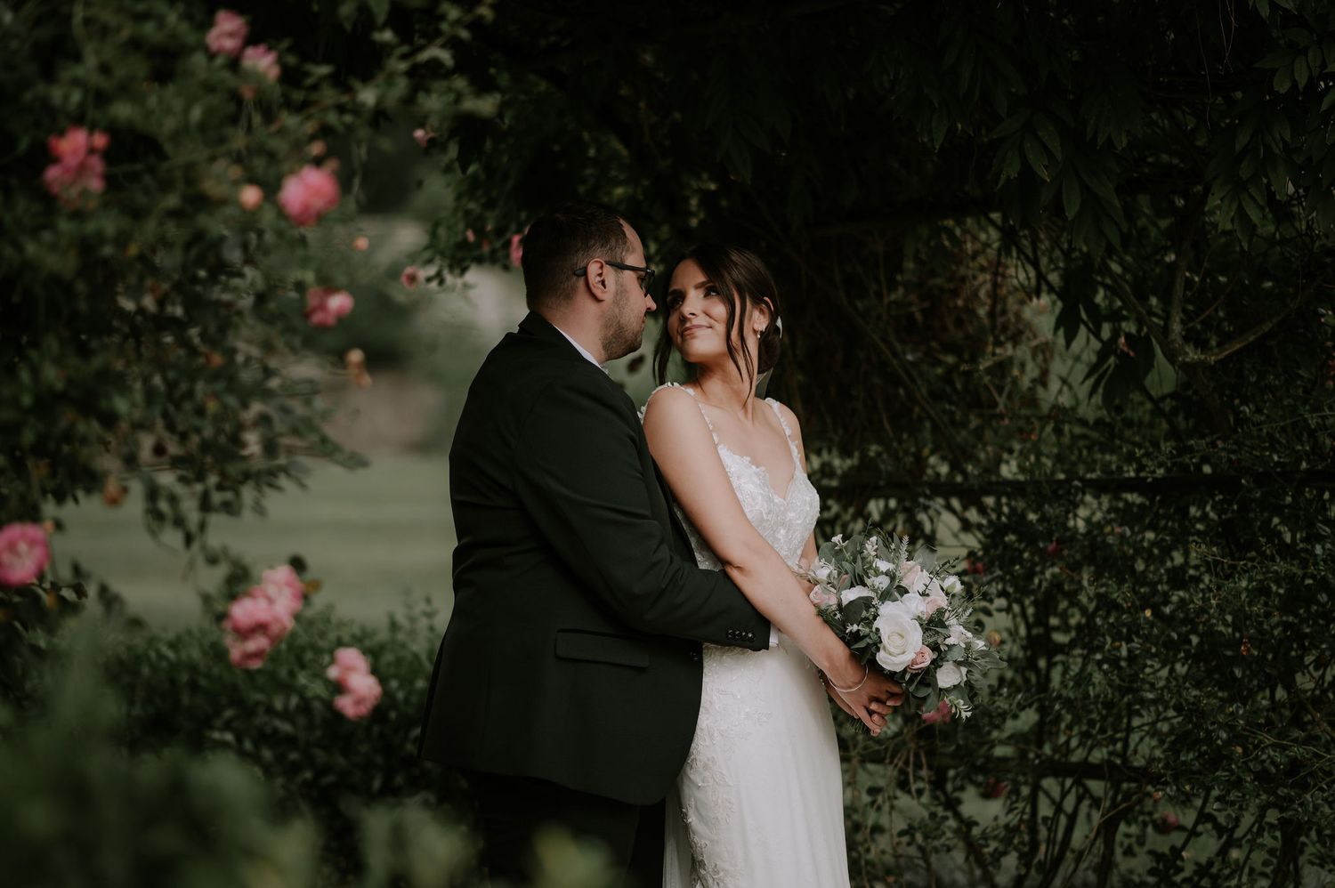 A bride looks back at her husband in a romantic embrace. Surrounded by pink roses and greenery. A melton Mowbray wedding