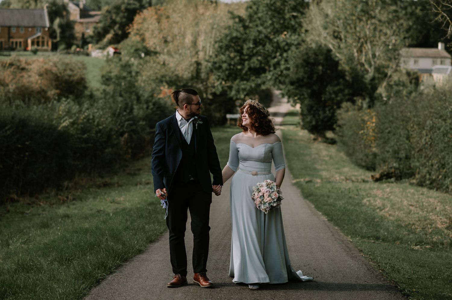 Couple walking hand in hand along a quiet country road bordered by open fields, smiling at each other.