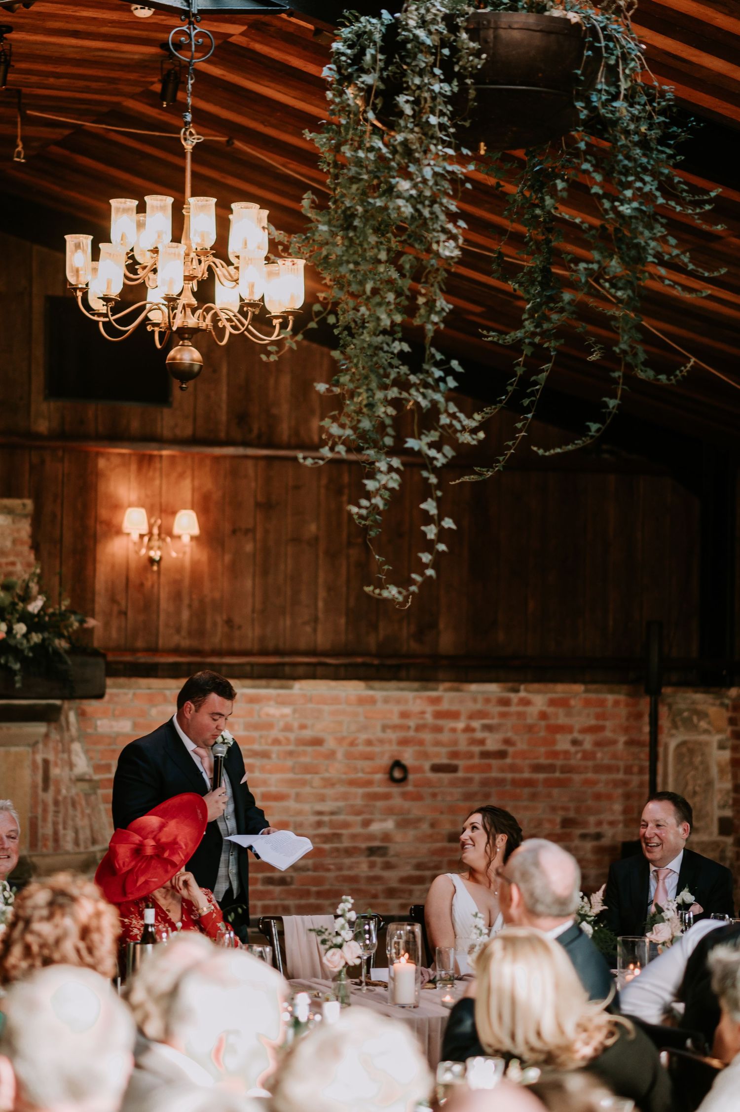 Wedding reception guests seated at long tables in a rustic barn venue with chandeliers and greenery decorations overhead.